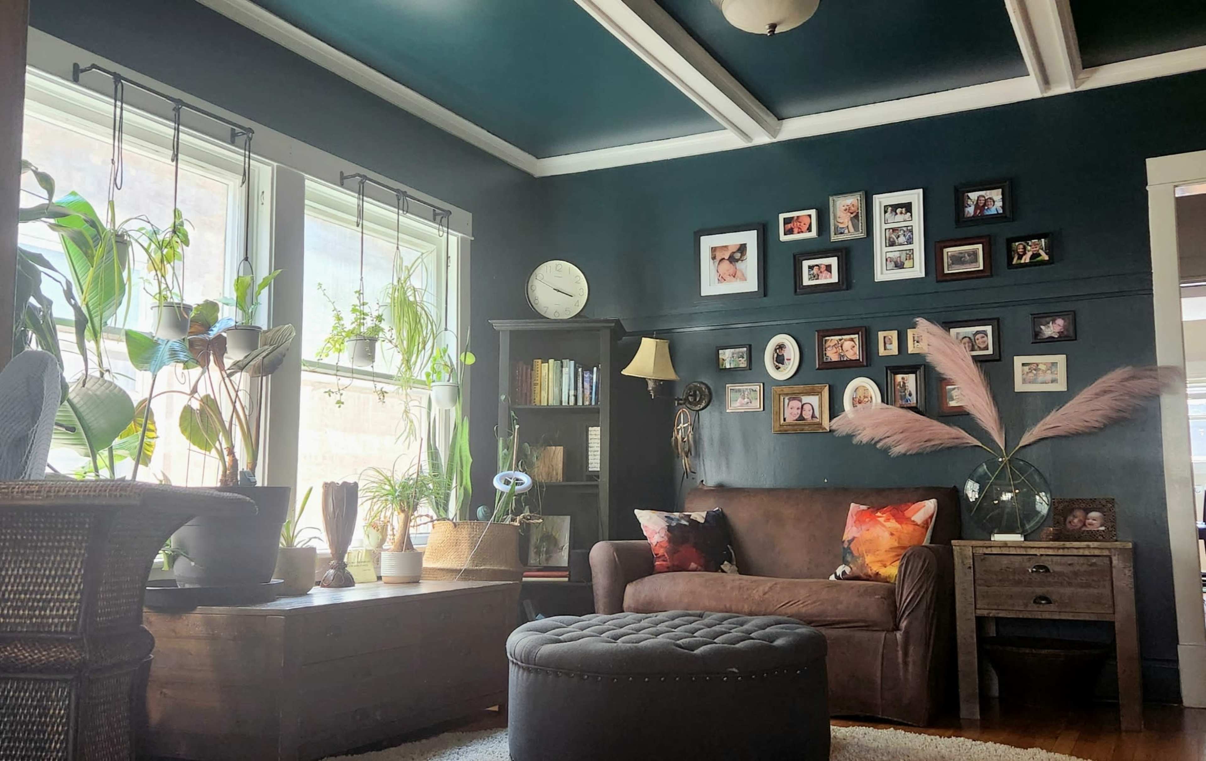 A living room features a dark blue wall with family photos, a brown sofa, potted plants near the windows, and a round ottoman in the center.