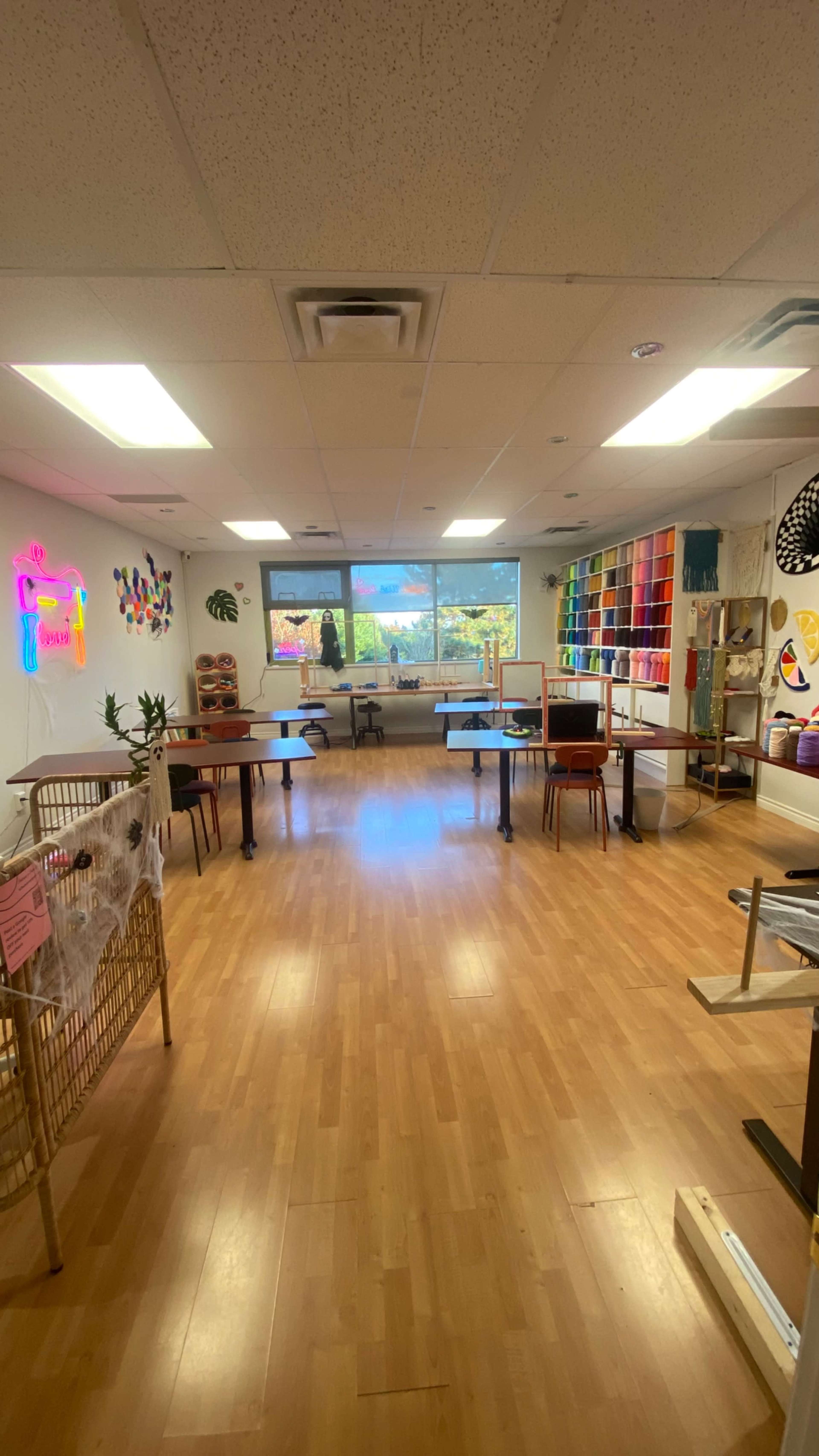A brightly lit craft studio features several tables, colorful wall displays, and a large window with a view of greenery outside.