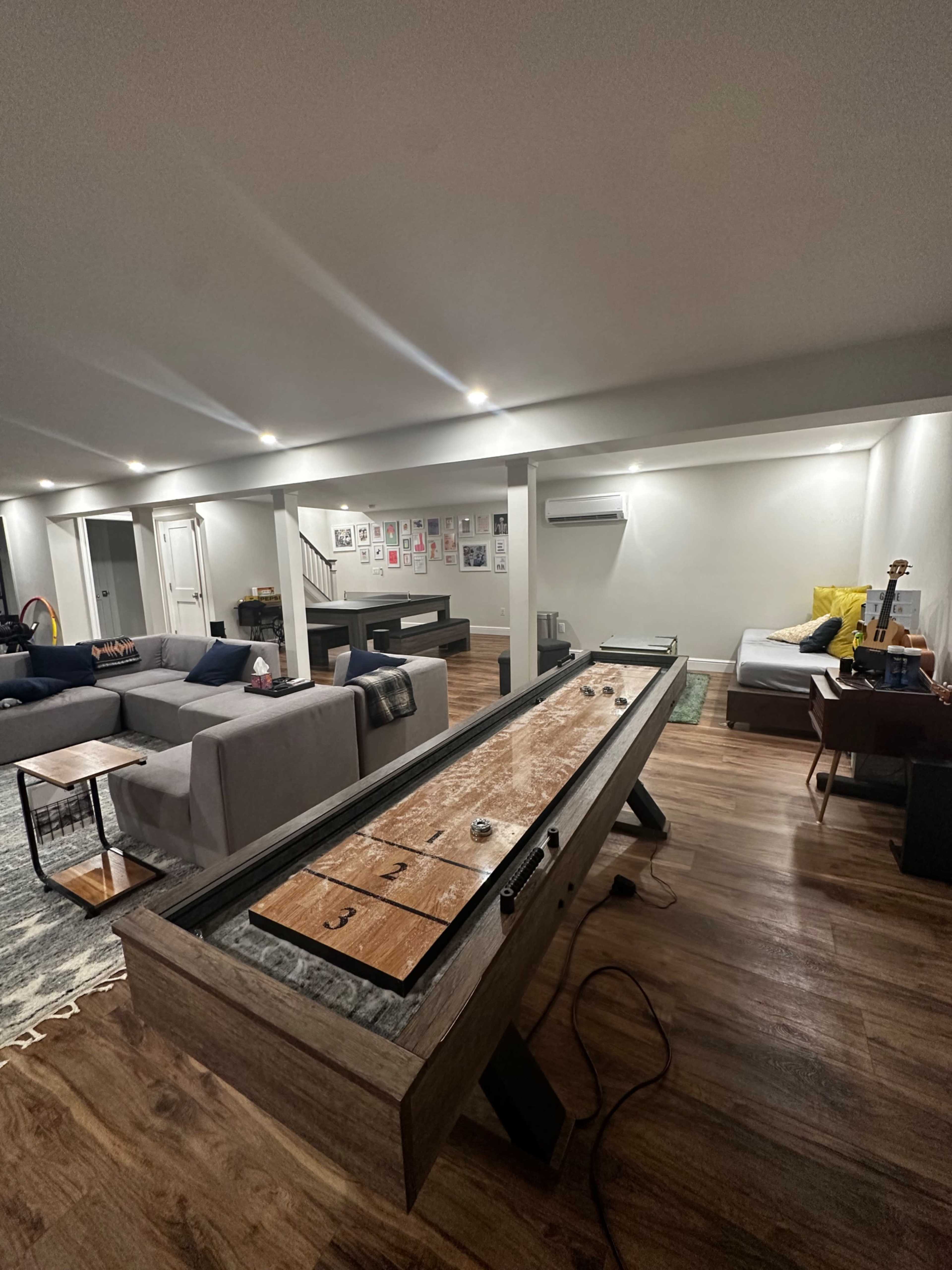 A shuffleboard table is positioned in the center of a modern, dimly lit basement lounge that features various seating areas and wall art.