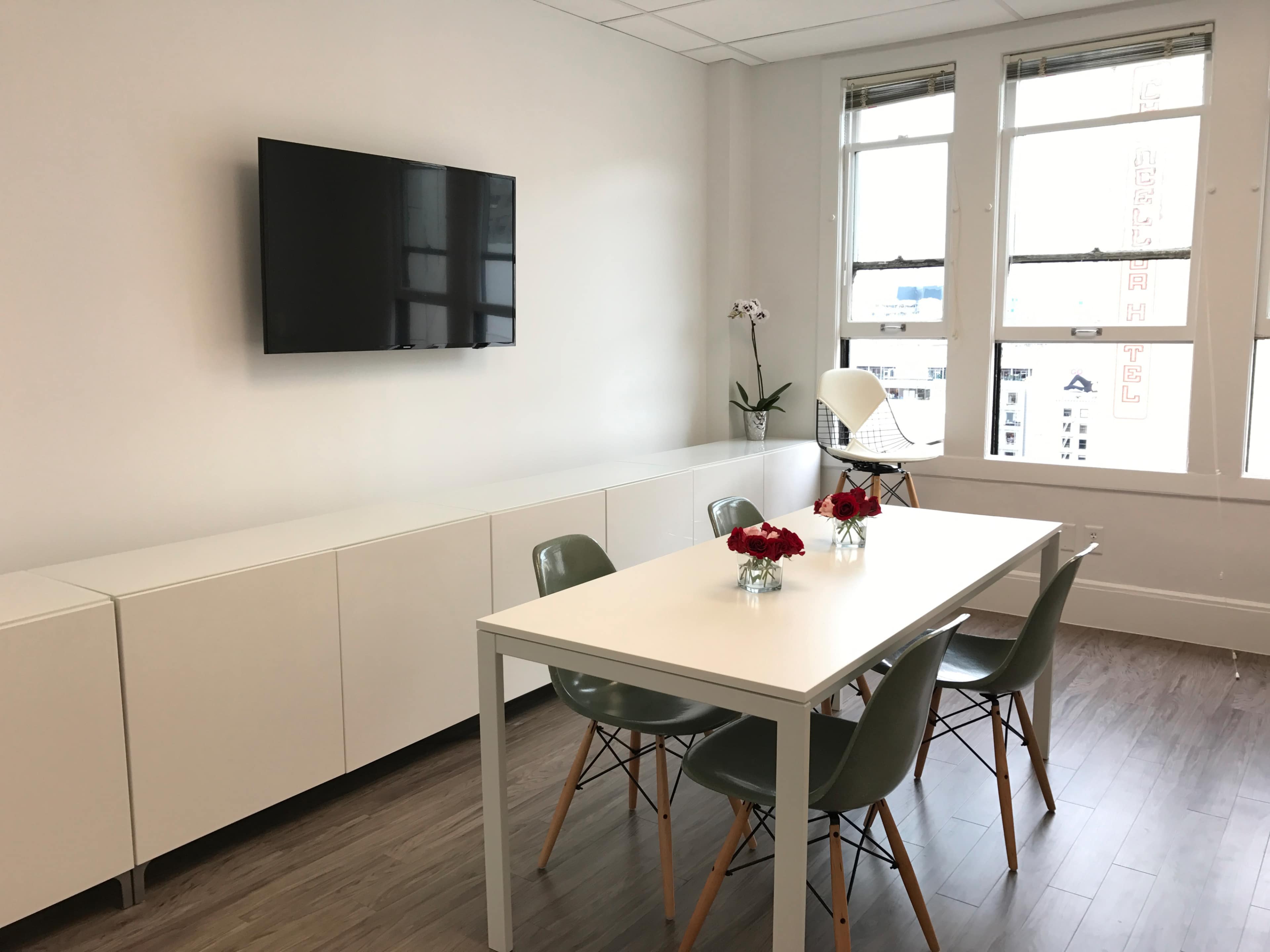 A modern conference room features a white table with four chairs, a television mounted on the wall, and large windows letting in natural light.