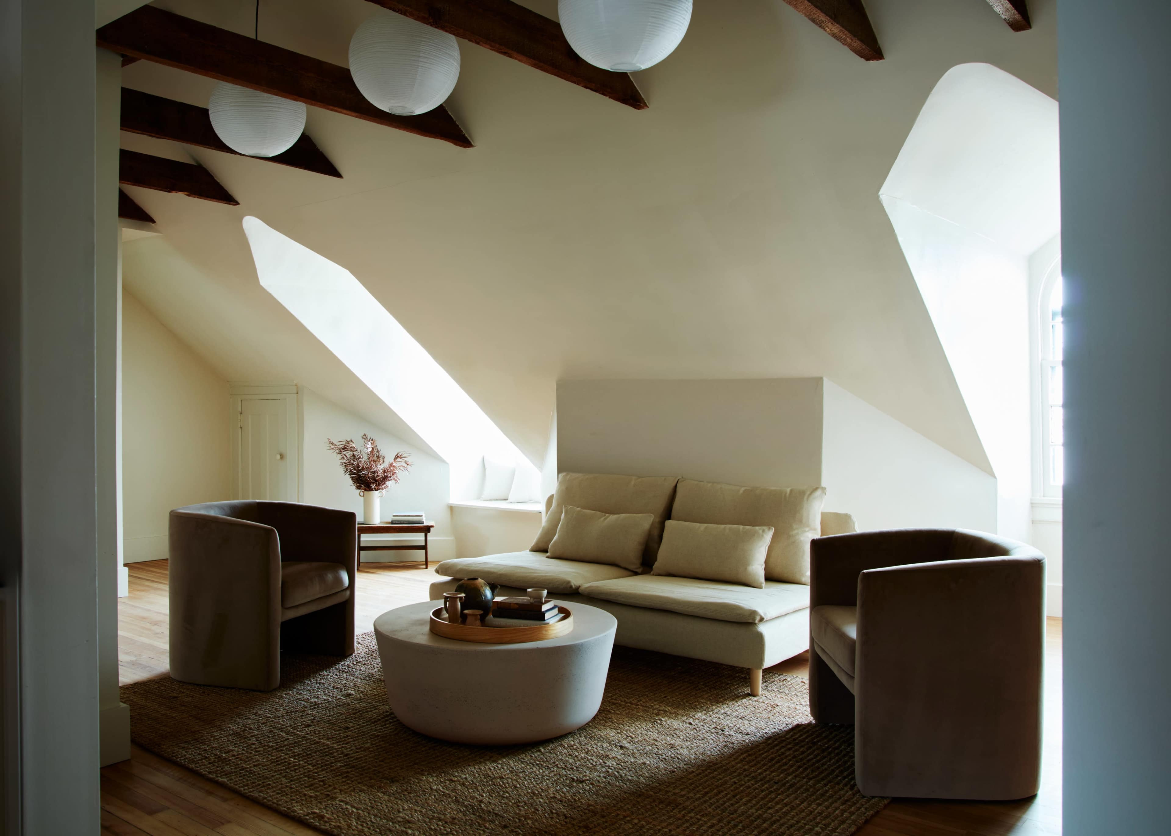 A spacious living room features a modern sofa, two armchairs, and a round coffee table, all set against slanted ceilings and large windows.