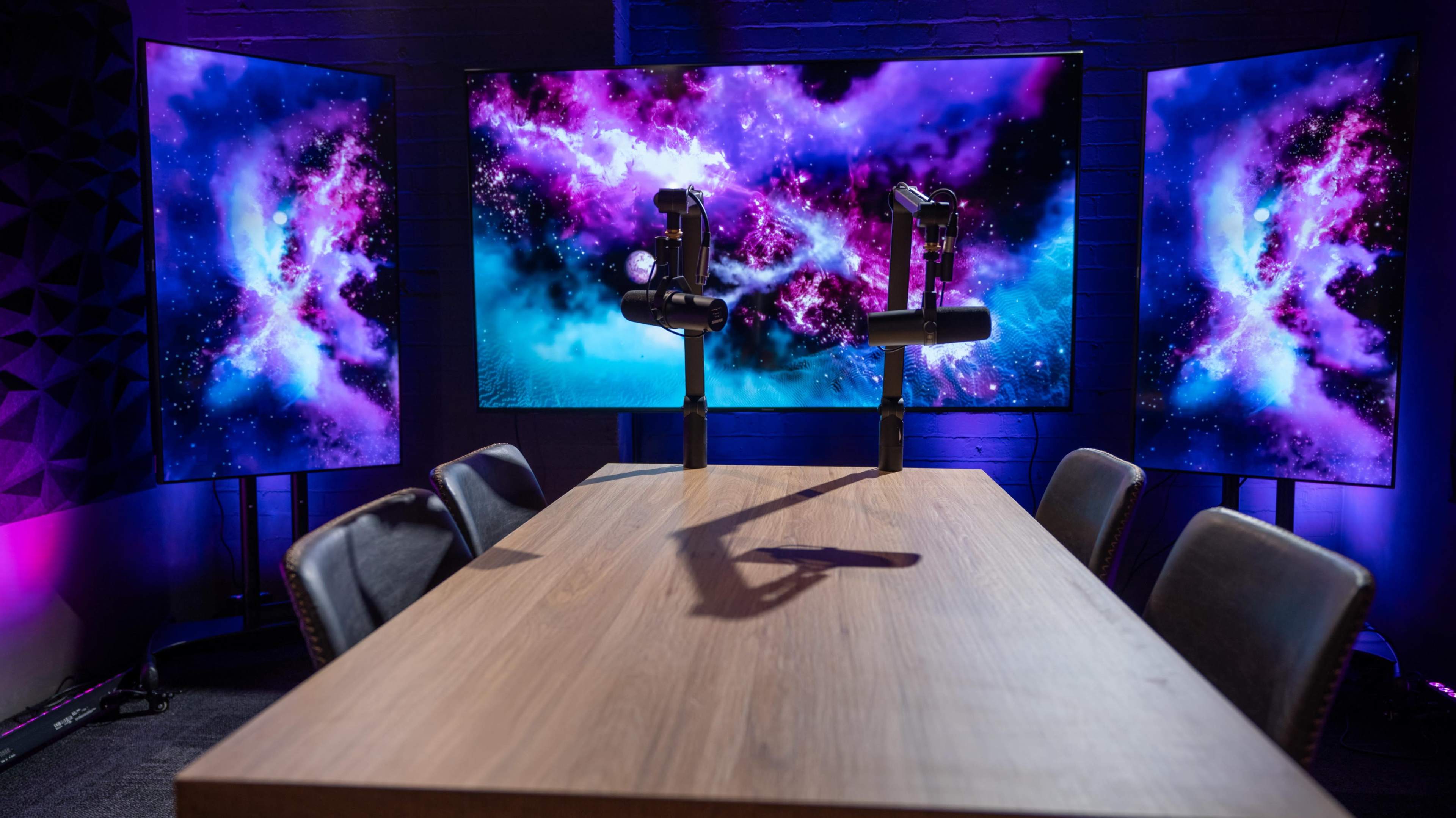 A wooden conference table is positioned in front of two large screens displaying colorful cosmic visuals in a dimly lit room.