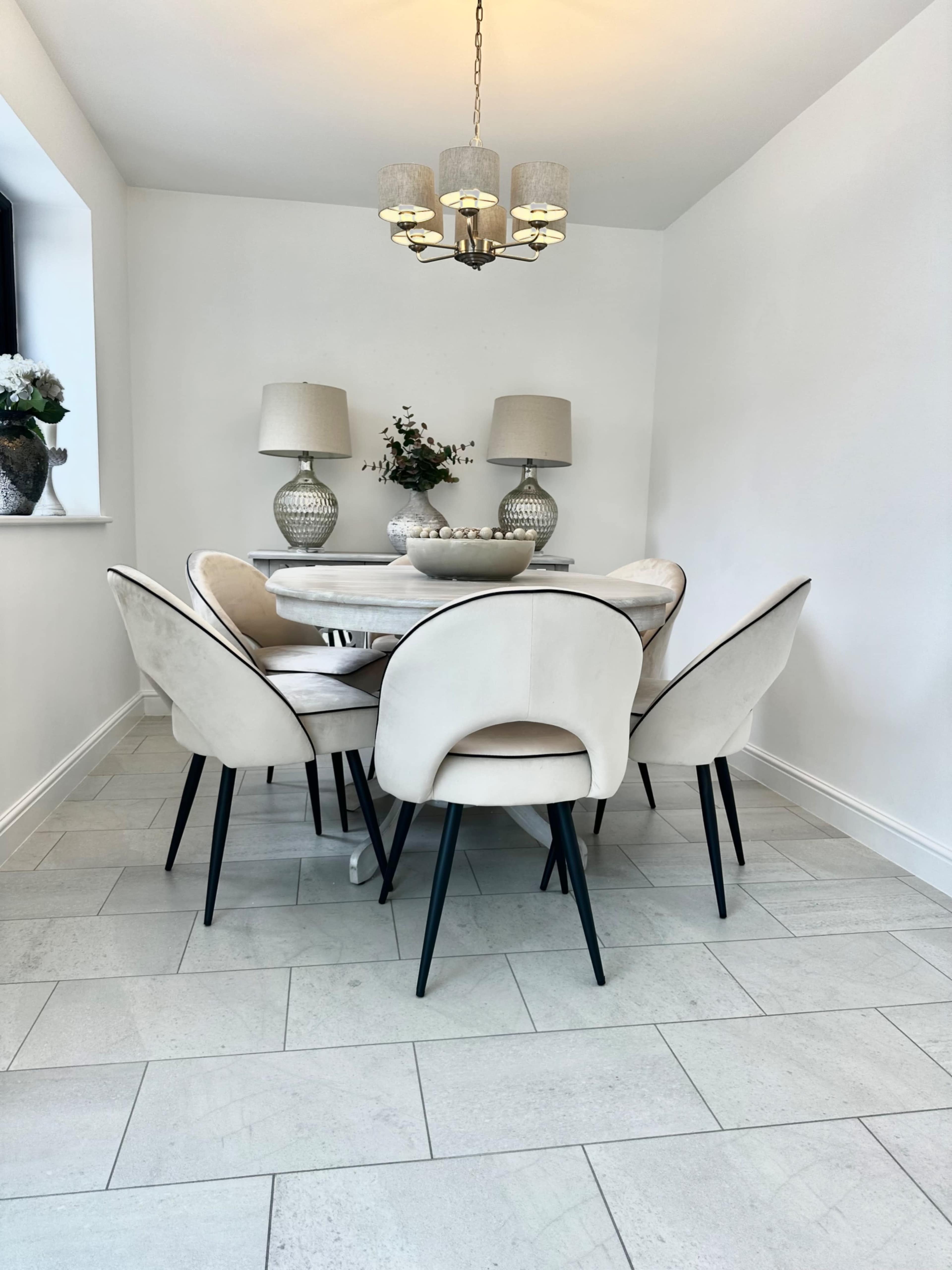 A round dining table surrounded by six cushioned chairs sits under a modern chandelier in a light-colored room with minimal decor.