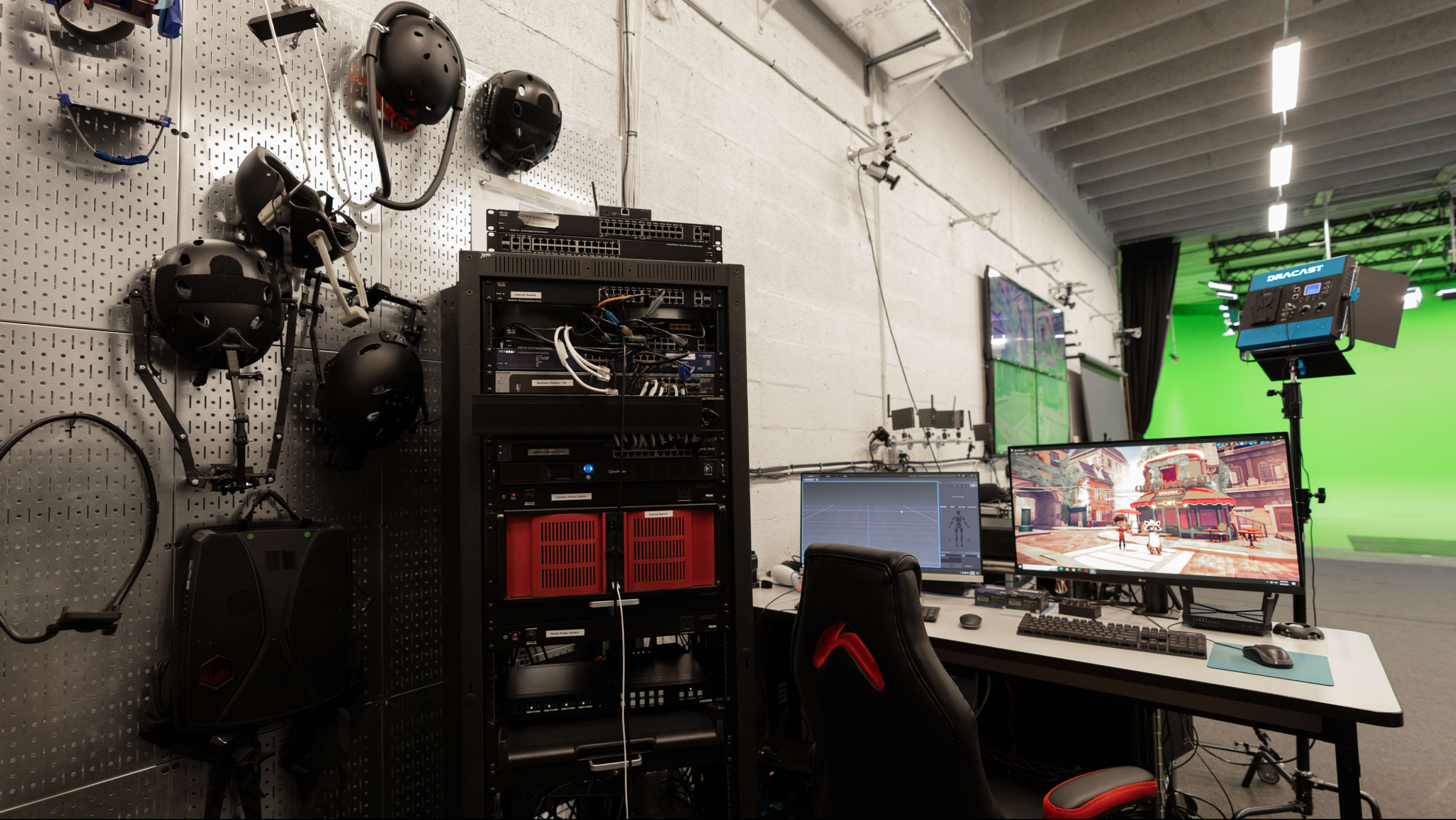 The image shows a studio setup with a computer workstation, a rack of equipment, and various helmets mounted on the wall.