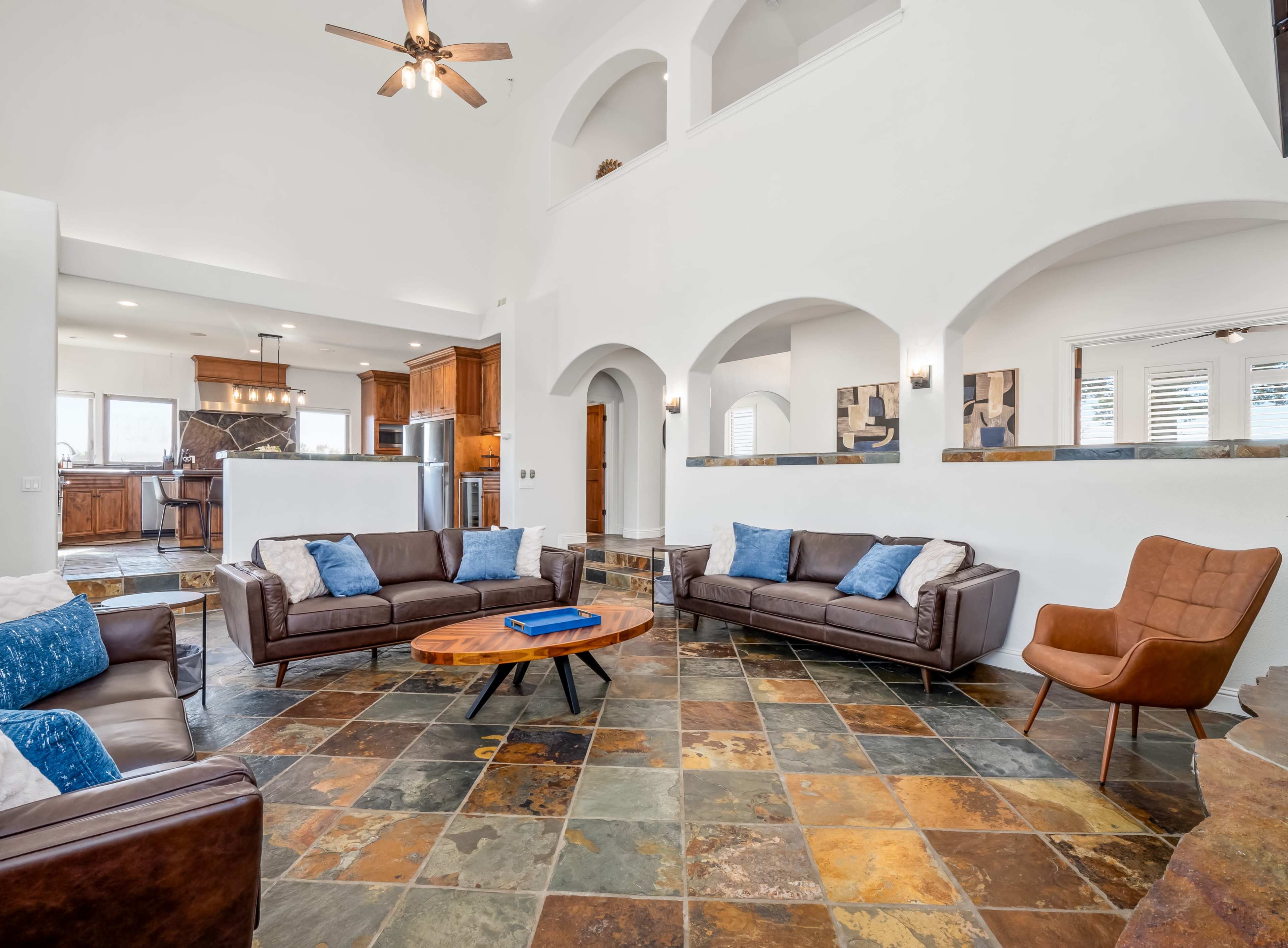 A spacious living area with two brown leather sofas, an orange armchair, a wooden coffee table, and a tiled floor, all set against white walls and arched openings.