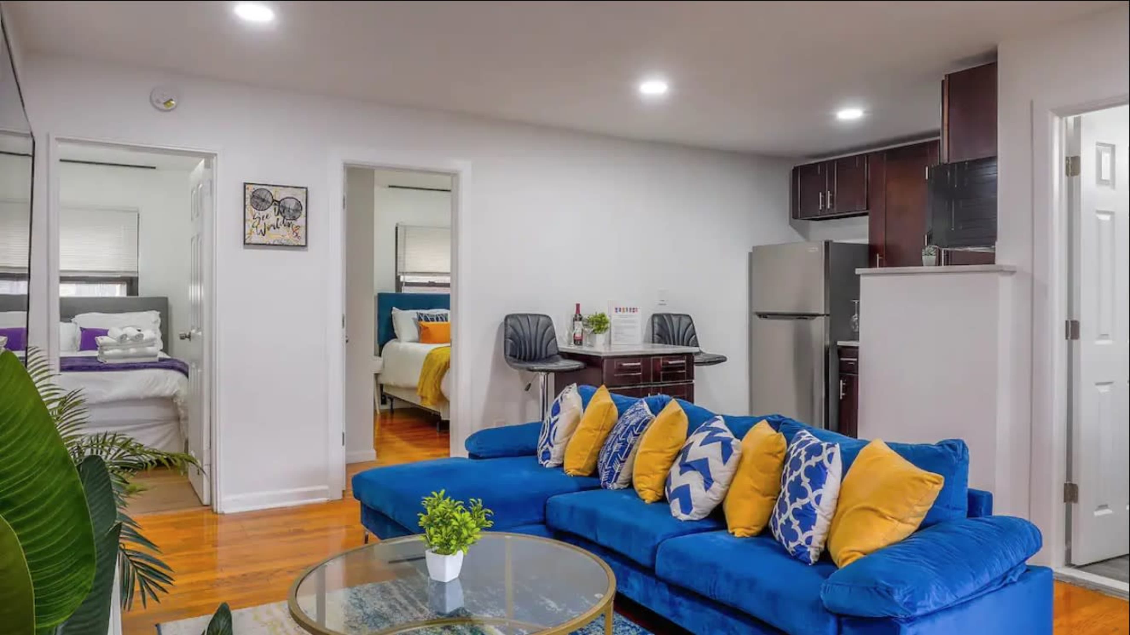 The image shows a modern living room with a blue sofa adorned with yellow and white pillows, a glass coffee table, and a view into a kitchen and adjacent bedrooms.