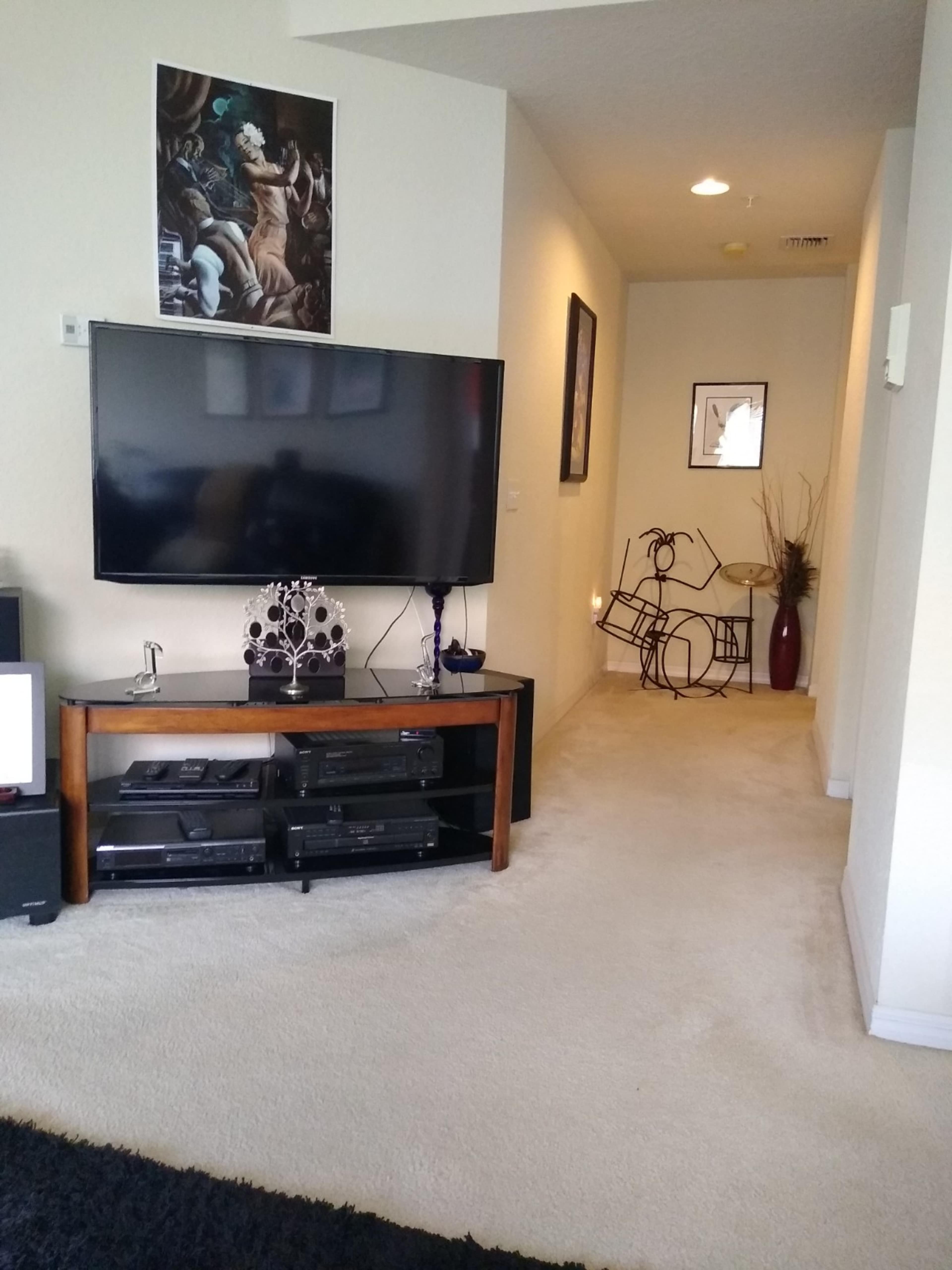 The scene features a living room with a large television on a wooden stand, leading to a hallway adorned with wall art and a decorative vase.
