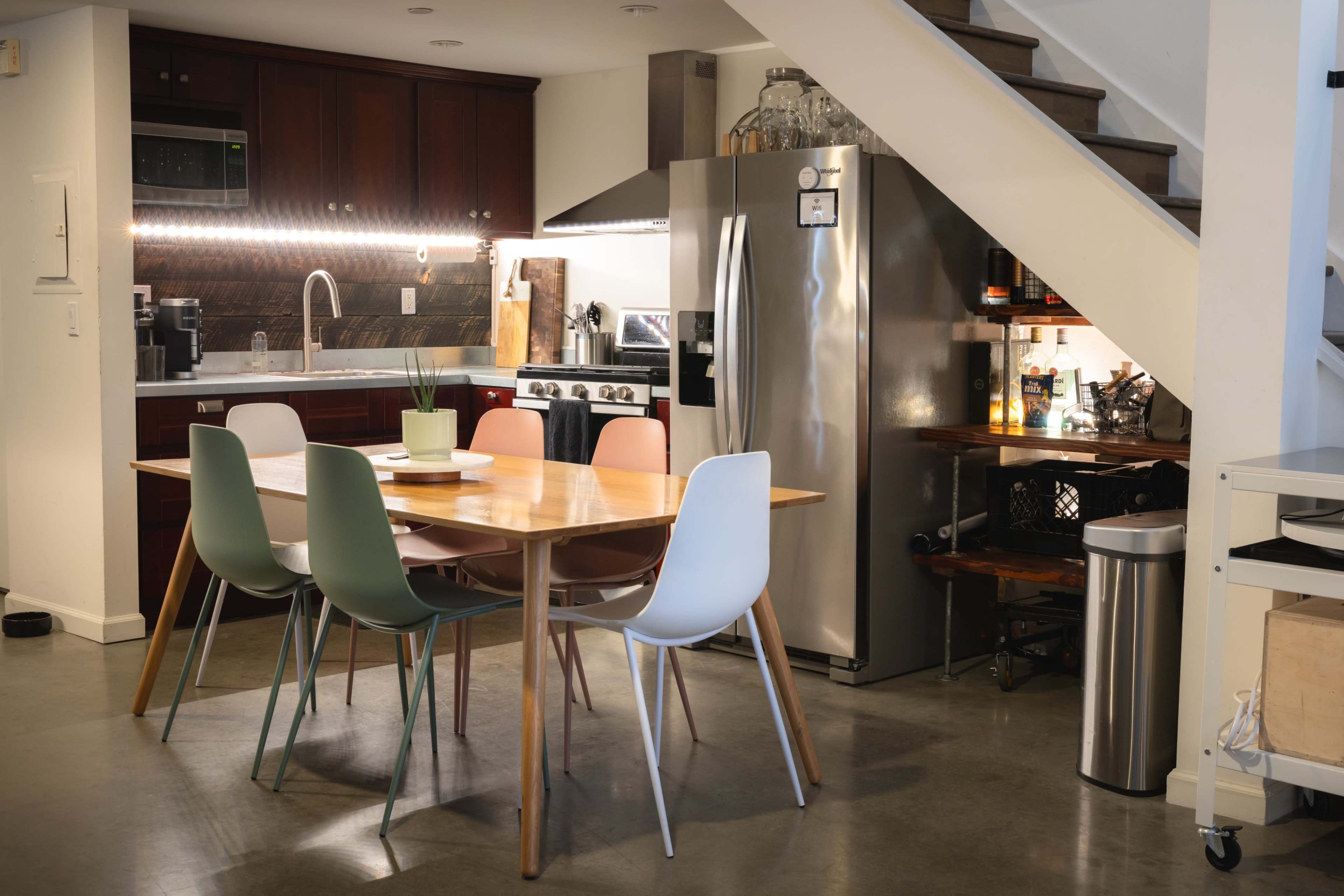 A modern kitchen features a dining table with four chairs, stainless steel appliances, and dark wooden cabinetry underneath a staircase.