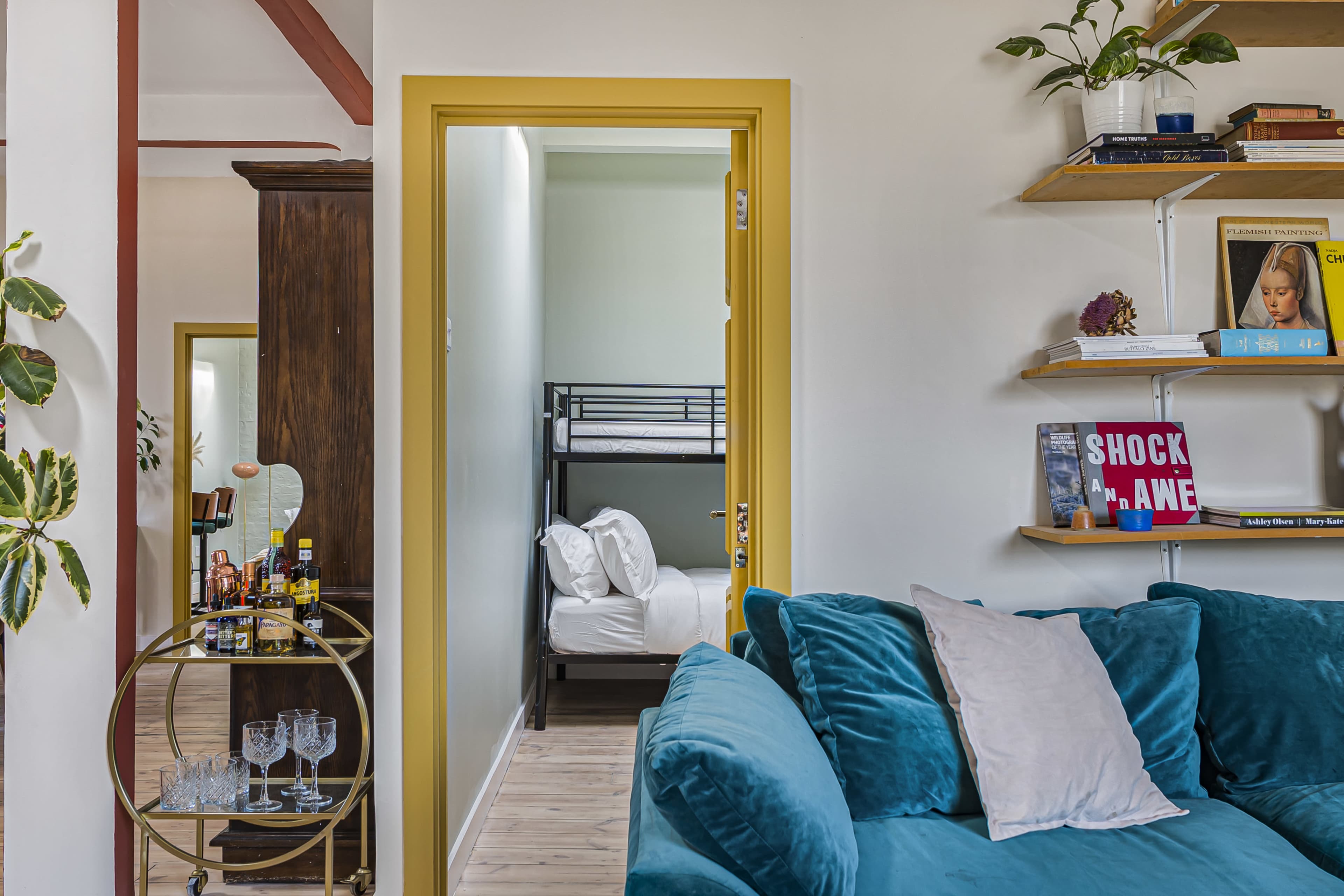 The image shows a living space with a teal couch, a small bar cart, and a doorway leading to a room with bunk beds.
