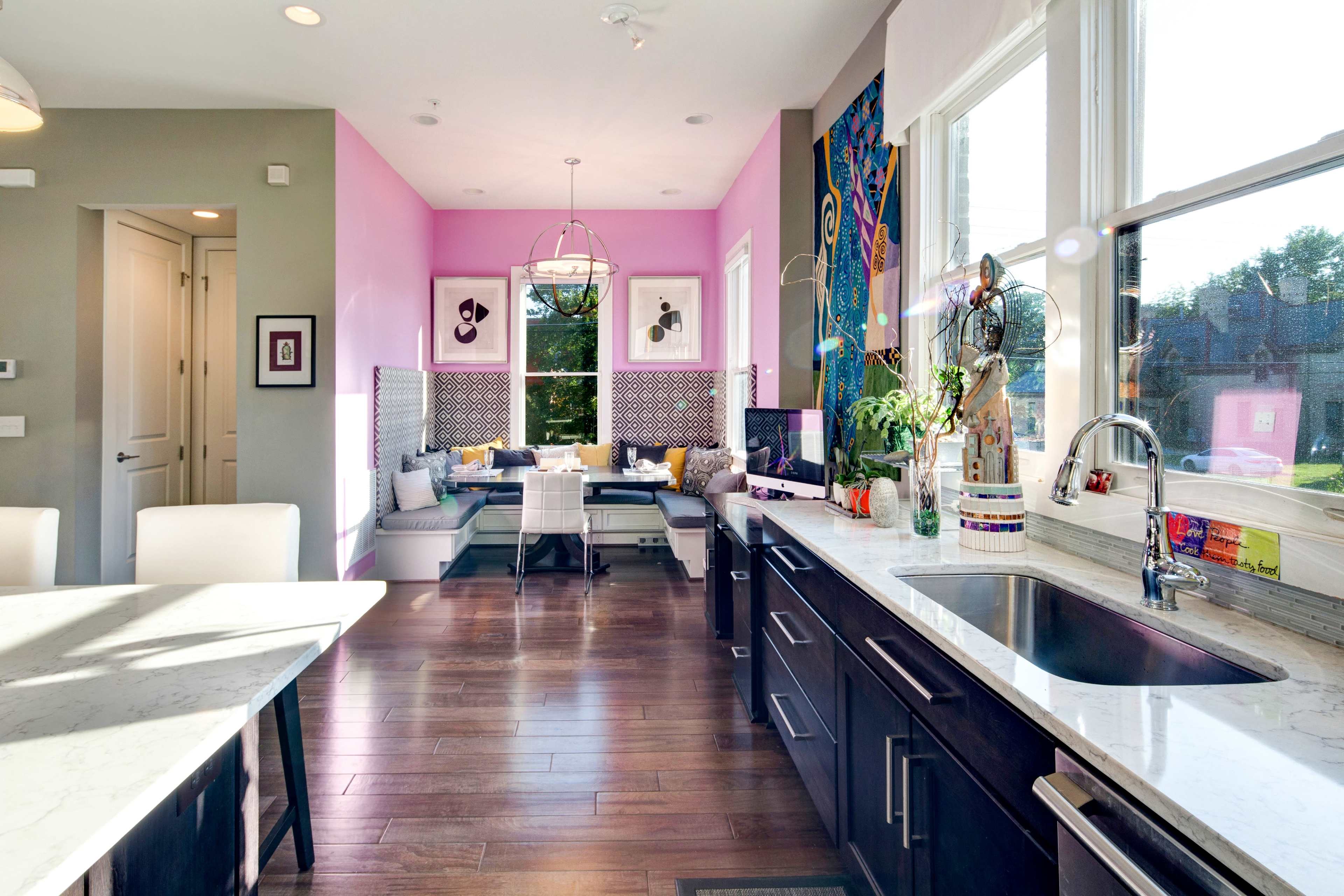 The image depicts a modern kitchen with a marble countertop, dark cabinetry, and a dining area featuring a pink wall and a seating nook by large windows.
