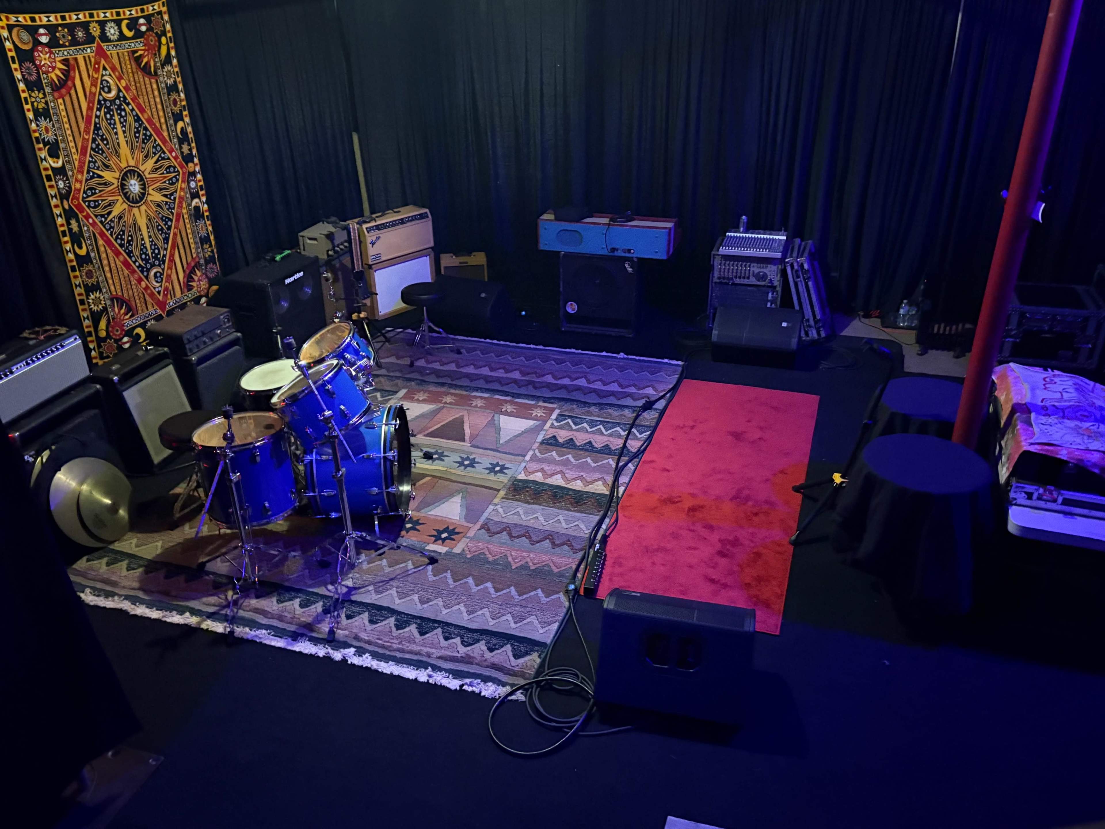 The image shows a music rehearsal space with a blue drum set, various amplifiers, a vintage keyboard, and a patterned rug on the floor, surrounded by black curtains.