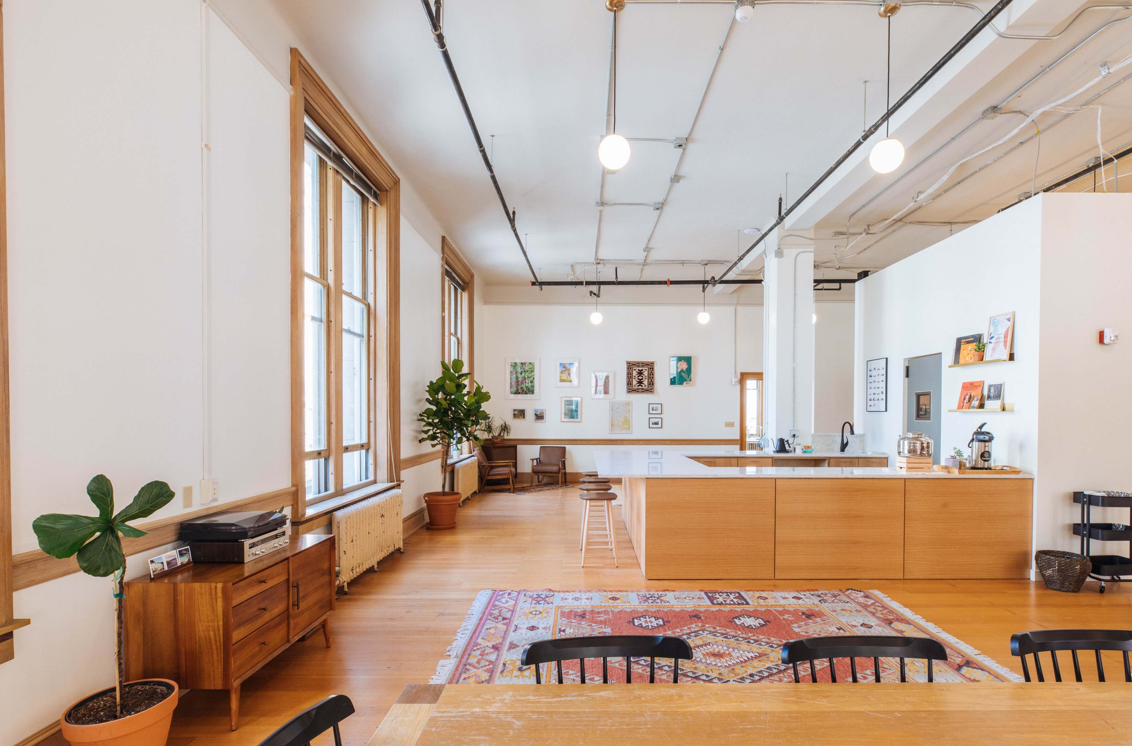 The image shows a spacious, well-lit kitchen and dining area with wooden furniture, a large window, and various decorative items on the walls.
