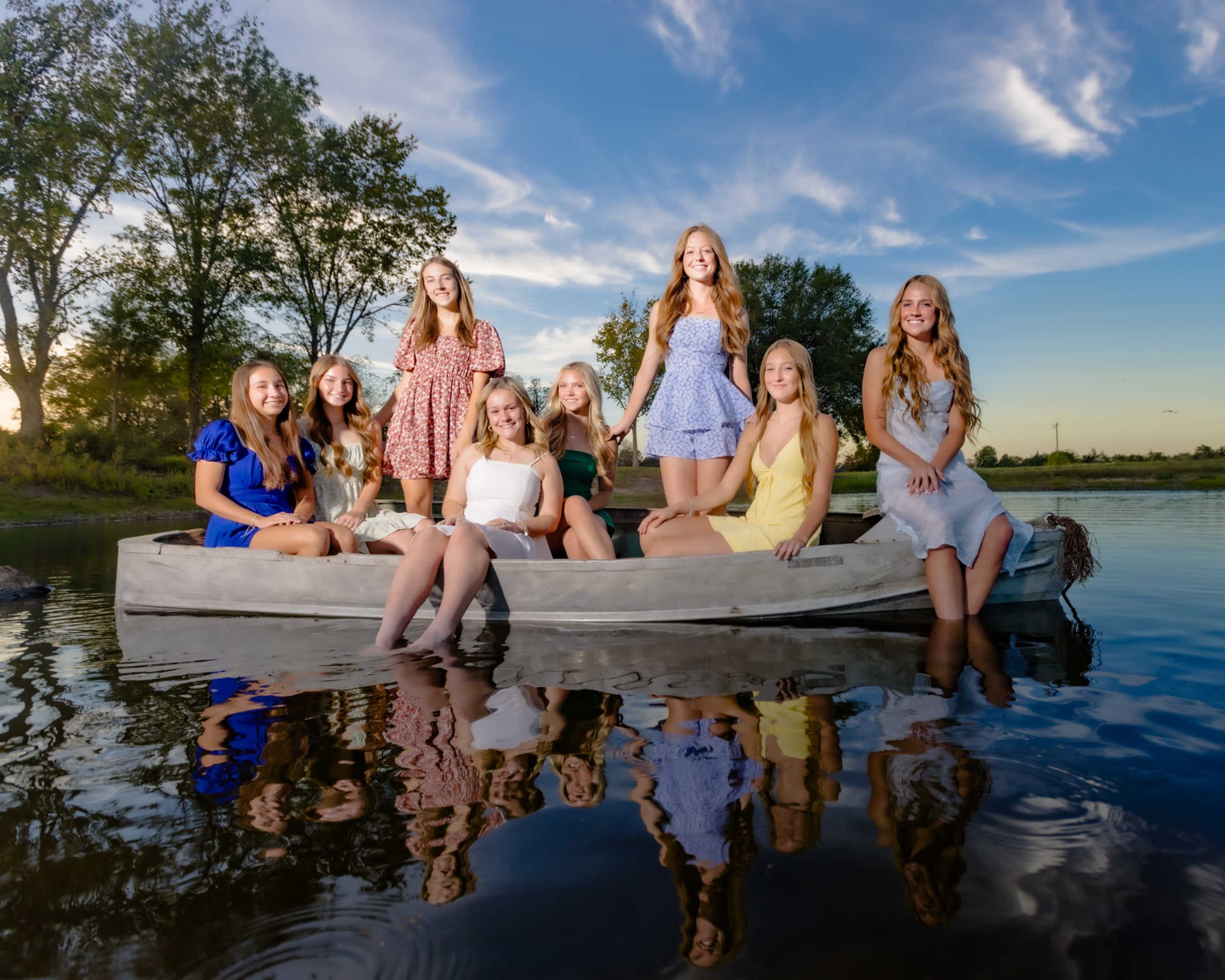 A group of nine young women in summer dresses stands in a boat on a calm lake, surrounded by trees and a blue sky with scattered clouds.
