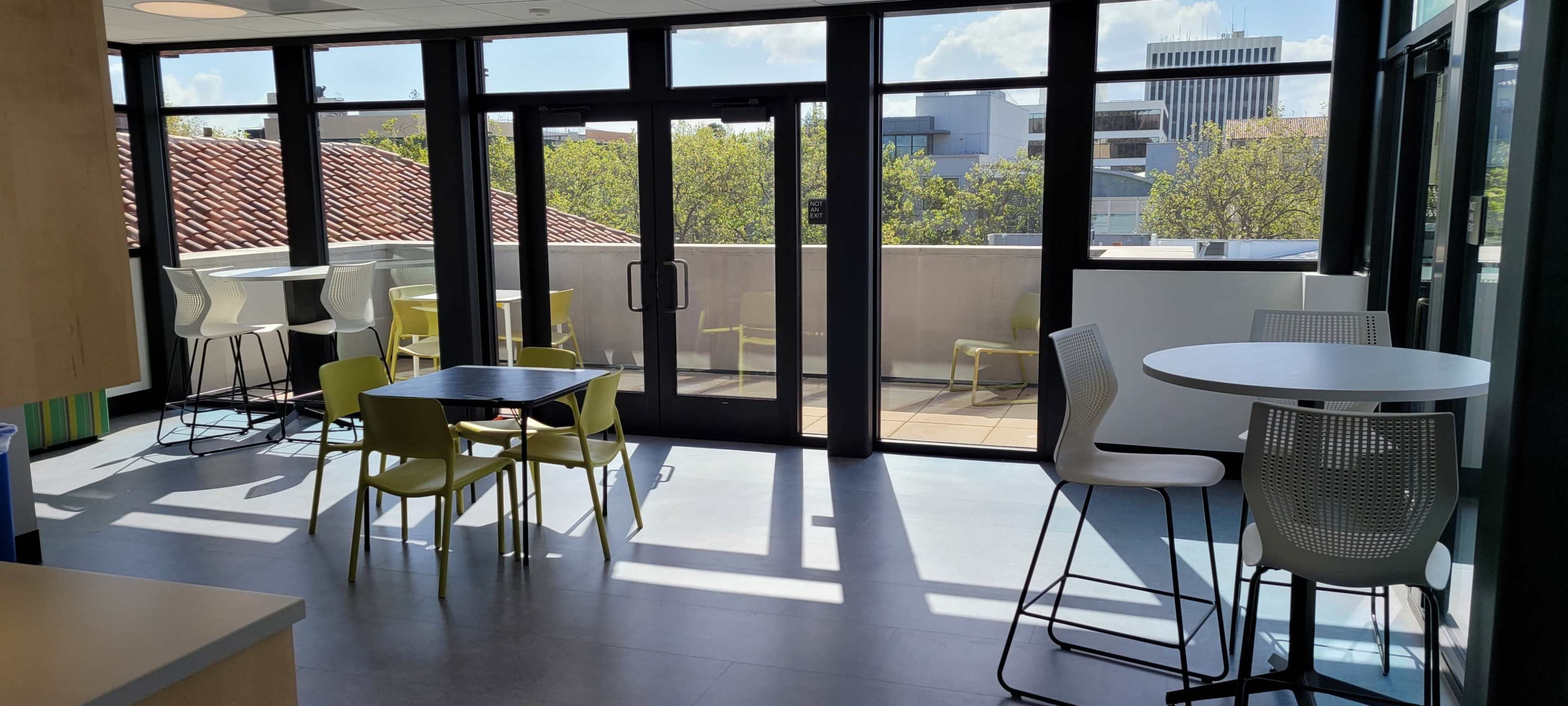 The image shows a brightly lit room with modern furniture, large windows, and a terrace view featuring tables and chairs.