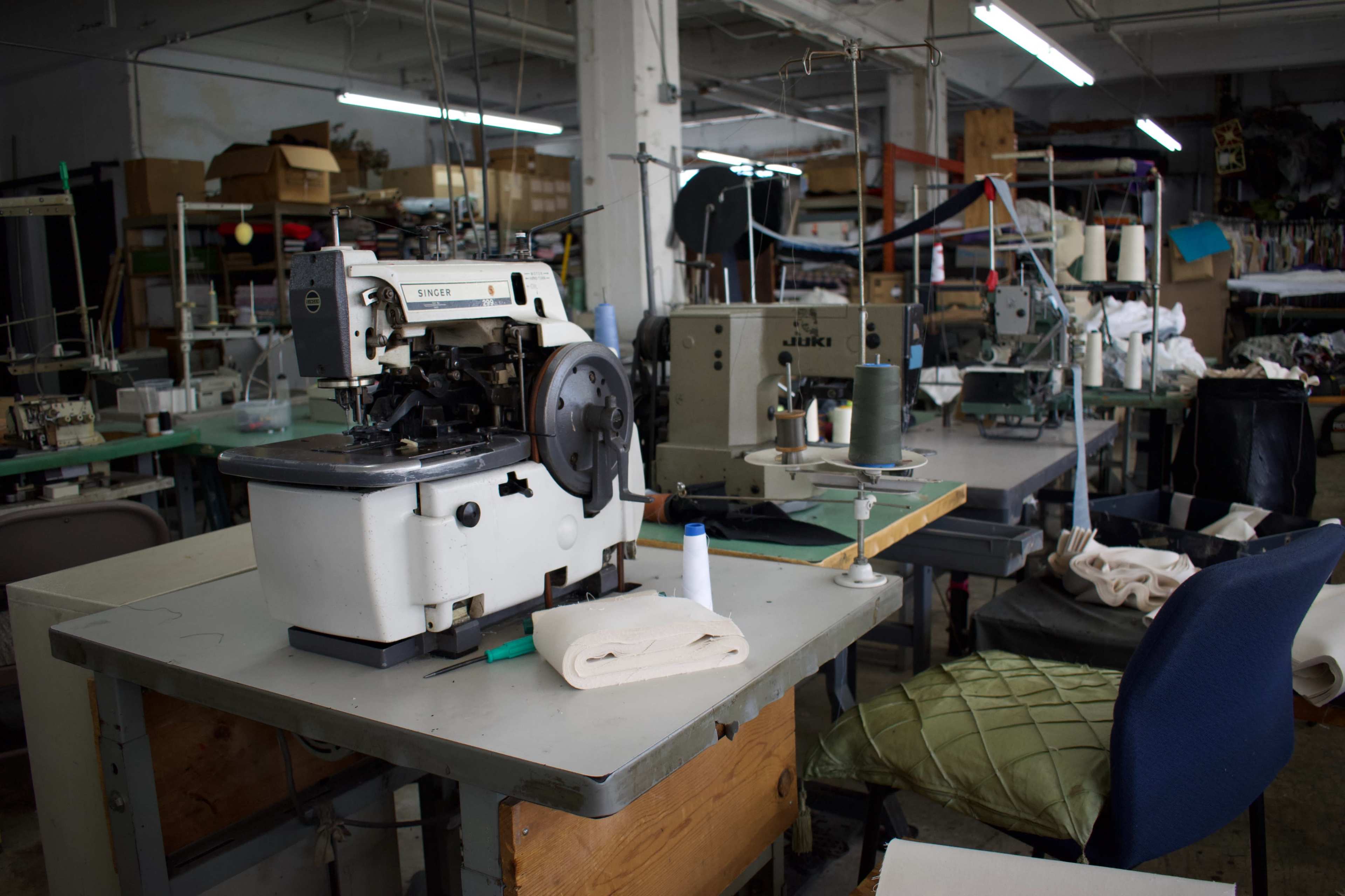 A textile workshop is filled with various sewing machines and cluttered tables, showcasing fabric, thread, and tools.