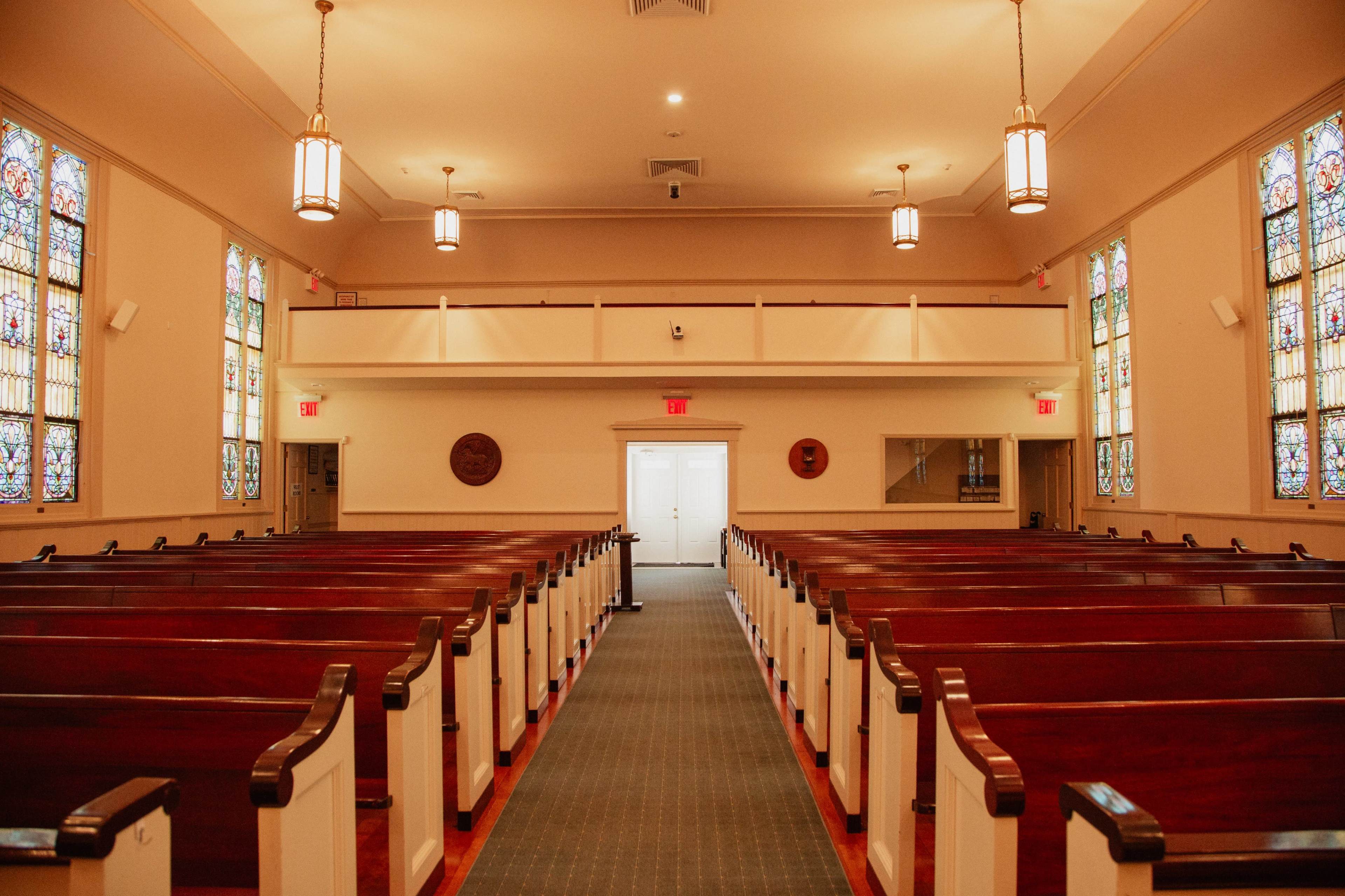The image shows the interior of a church with wooden pews, stained glass windows, and a central aisle leading to double doors.