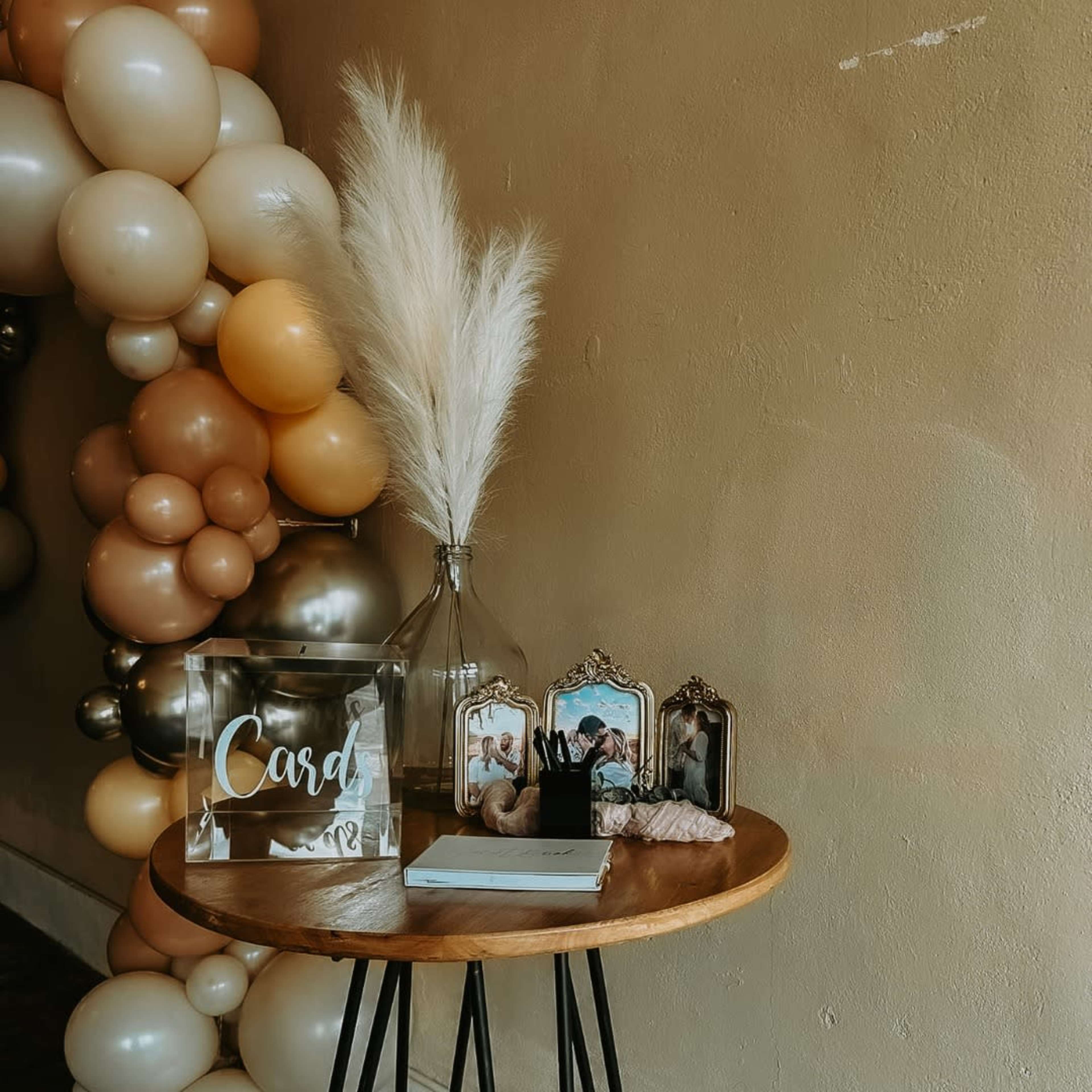 A wooden table displays a "Cards" sign, a vase with pampas grass, and framed photographs against a backdrop of balloons.