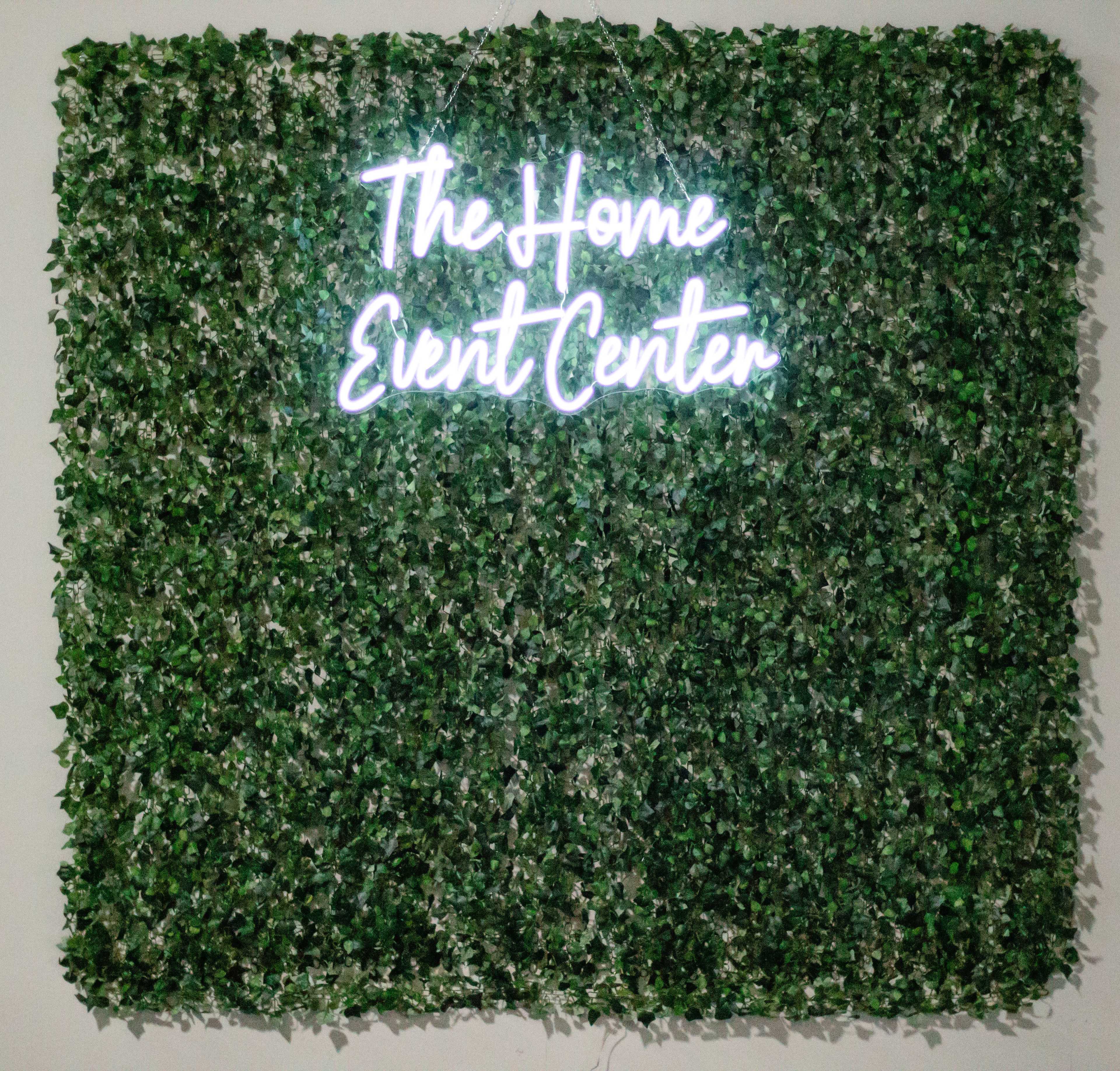 A neon sign reading "The Home Event Center" is displayed against a wall covered in artificial greenery.