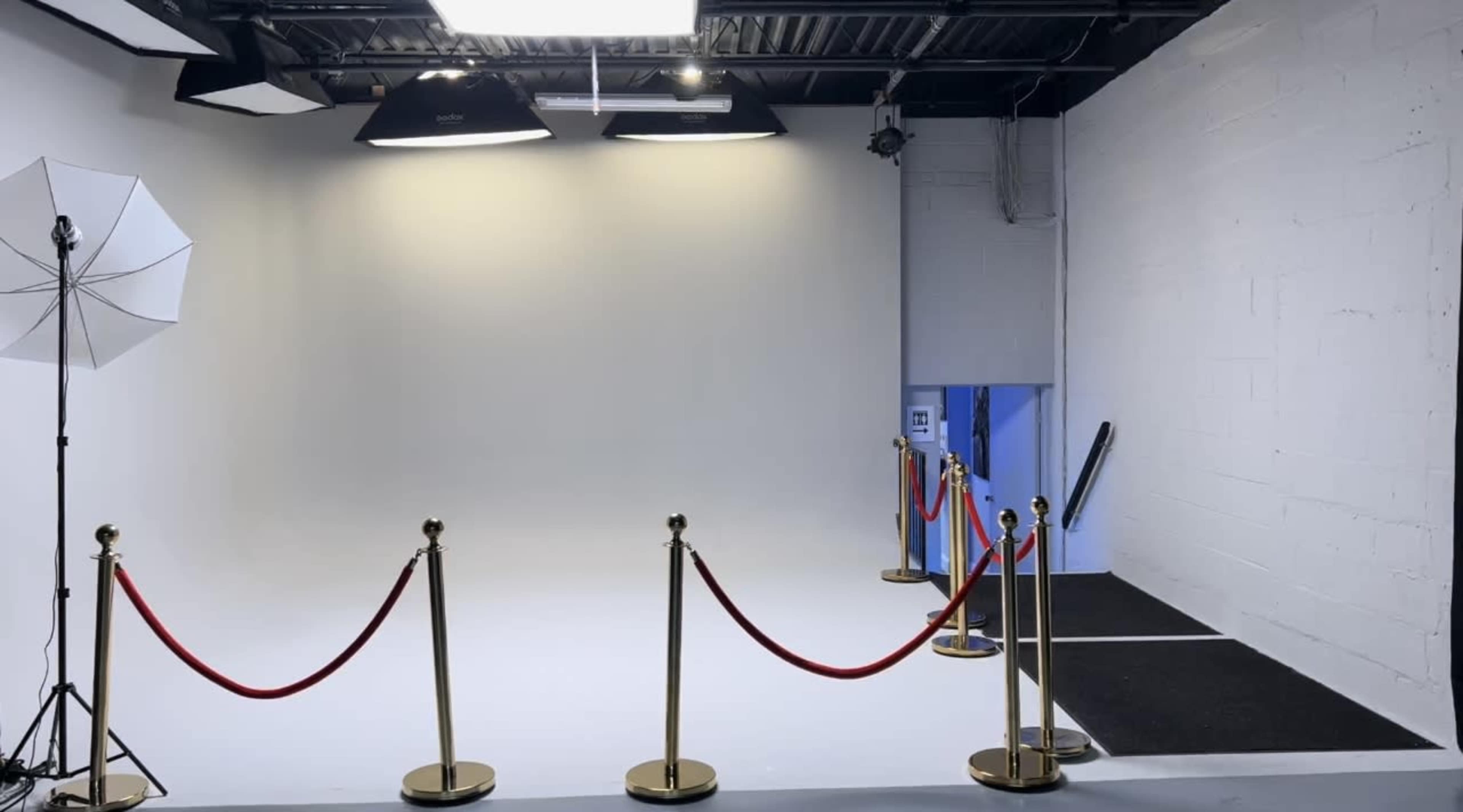 A photography studio features a white backdrop, a lighting setup, and a sectioned area with velvet ropes and stanchions.