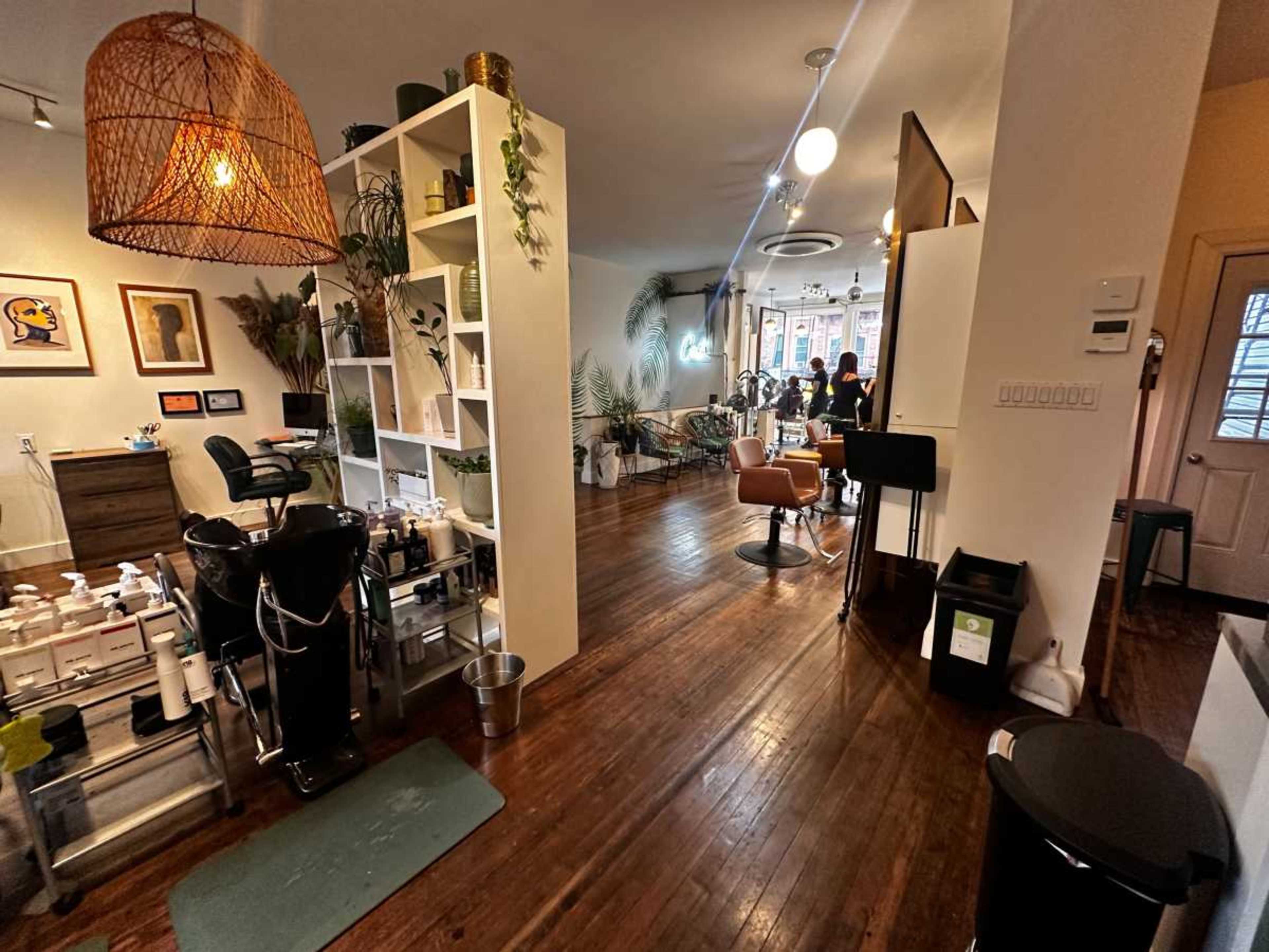 The image shows a brightly lit salon interior with wooden floors, styling stations, and decorative plants.