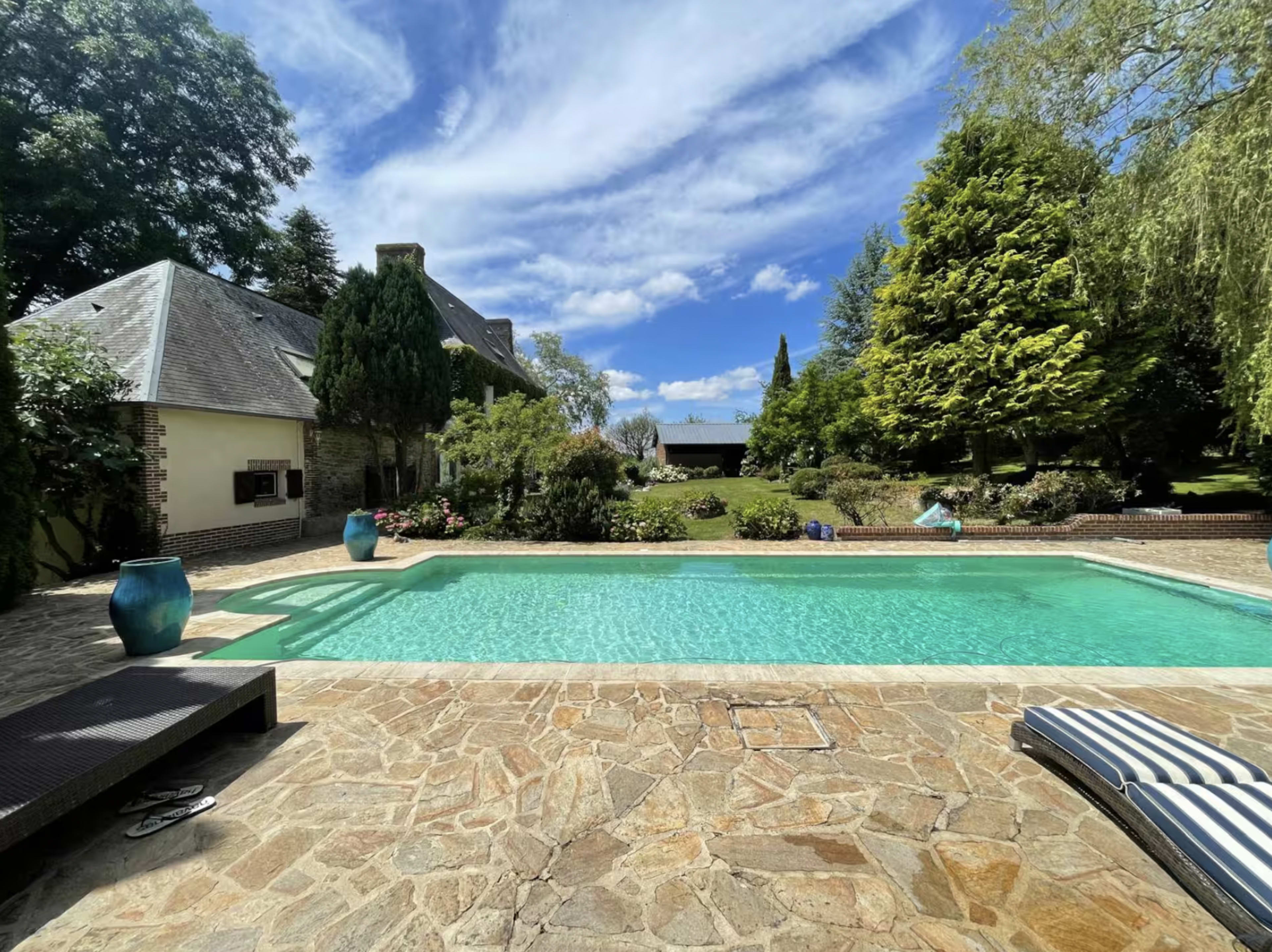 The image shows a clear swimming pool surrounded by stone pavers and lush greenery, with a house and blue sky in the background.