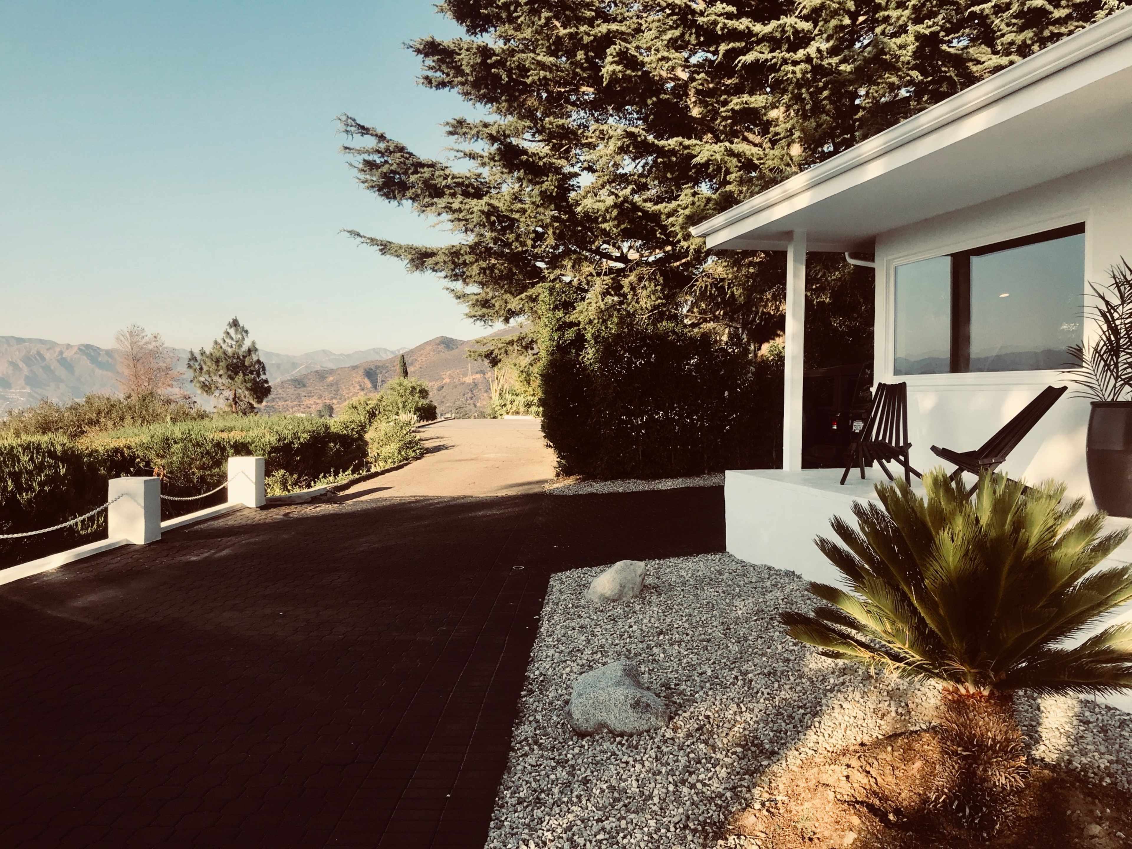 A modern white house with a patio overlooks a mountain landscape, bordered by neatly landscaped greenery and a paved driveway.