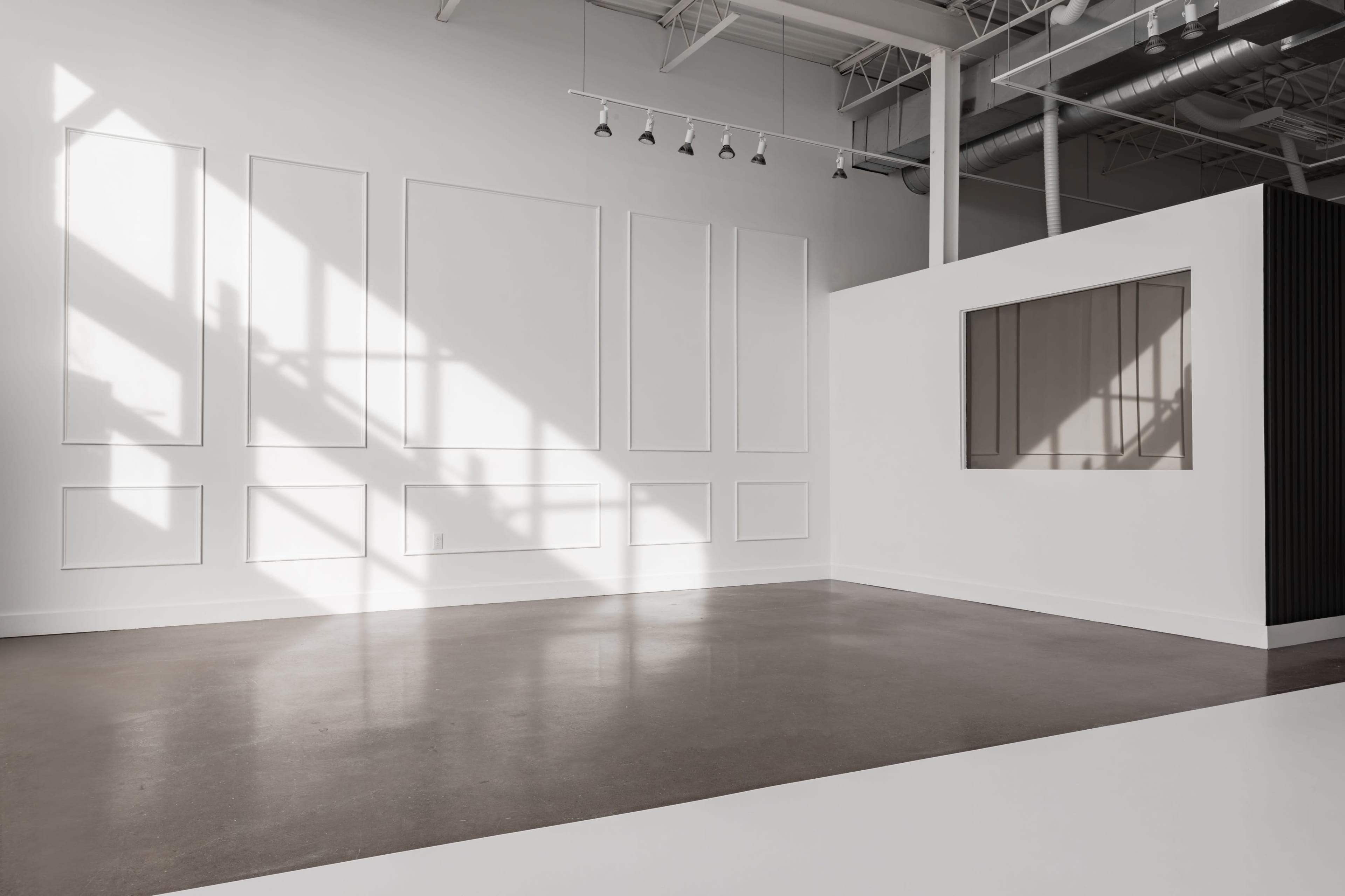 A spacious, minimally designed room with white walls, a large window casting shadows, and a feature wall with a window-like opening.