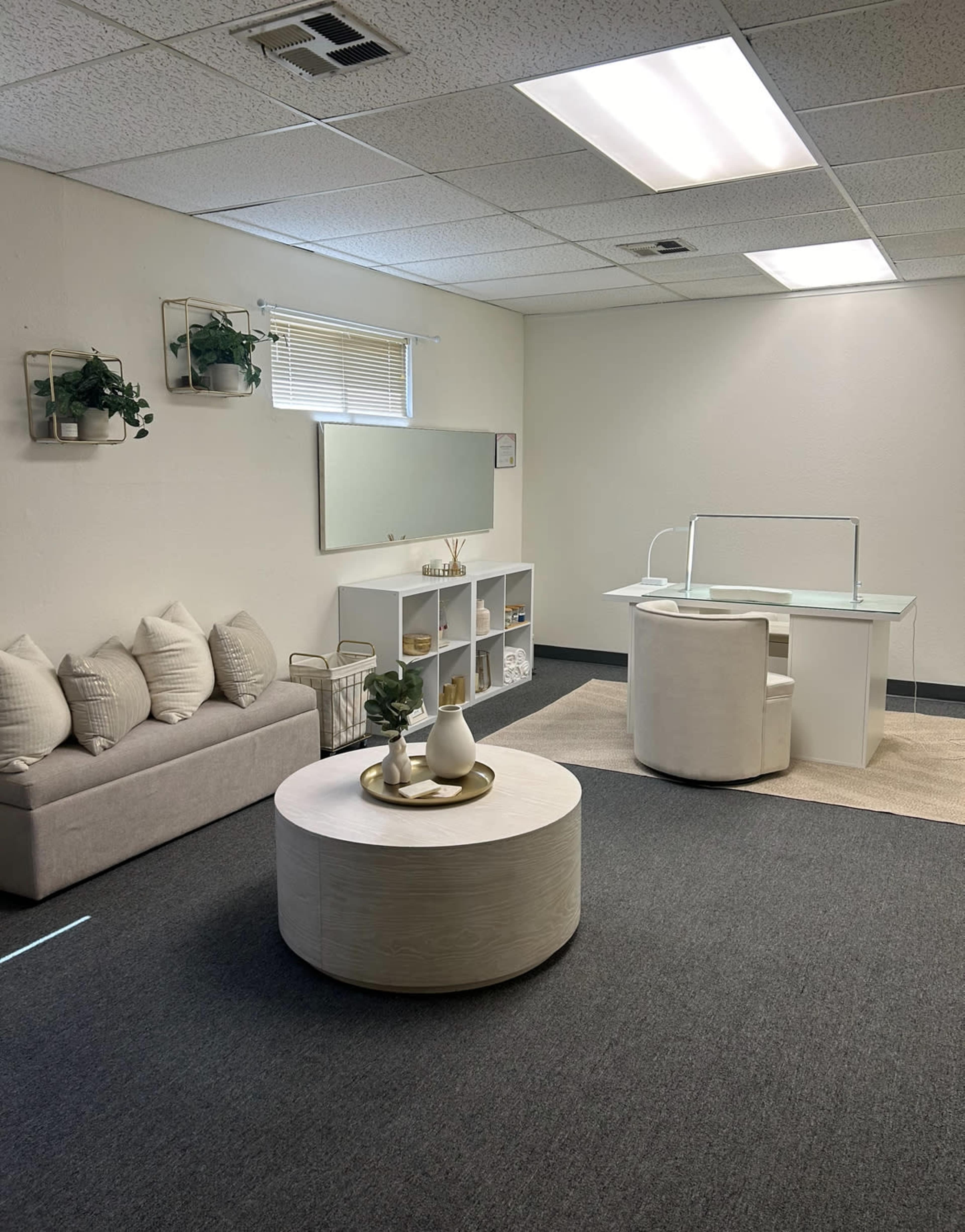 A minimalistic office space with a light-colored desk and chair, a round coffee table, and decorative plants against a neutral wall.