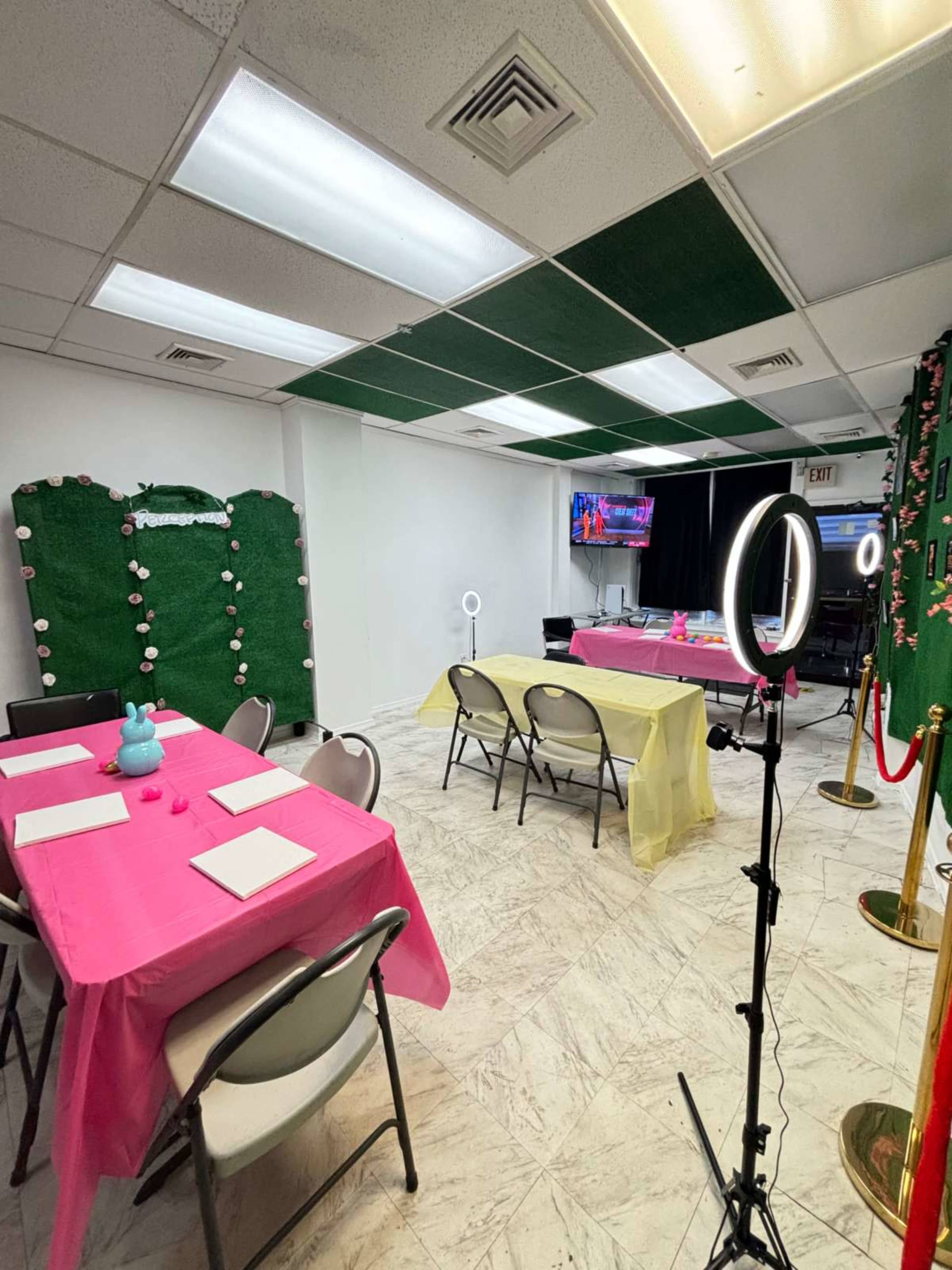 A brightly lit room set up for an event, featuring tables covered in pink and yellow tablecloths, with a green backdrop and a ring light positioned nearby.