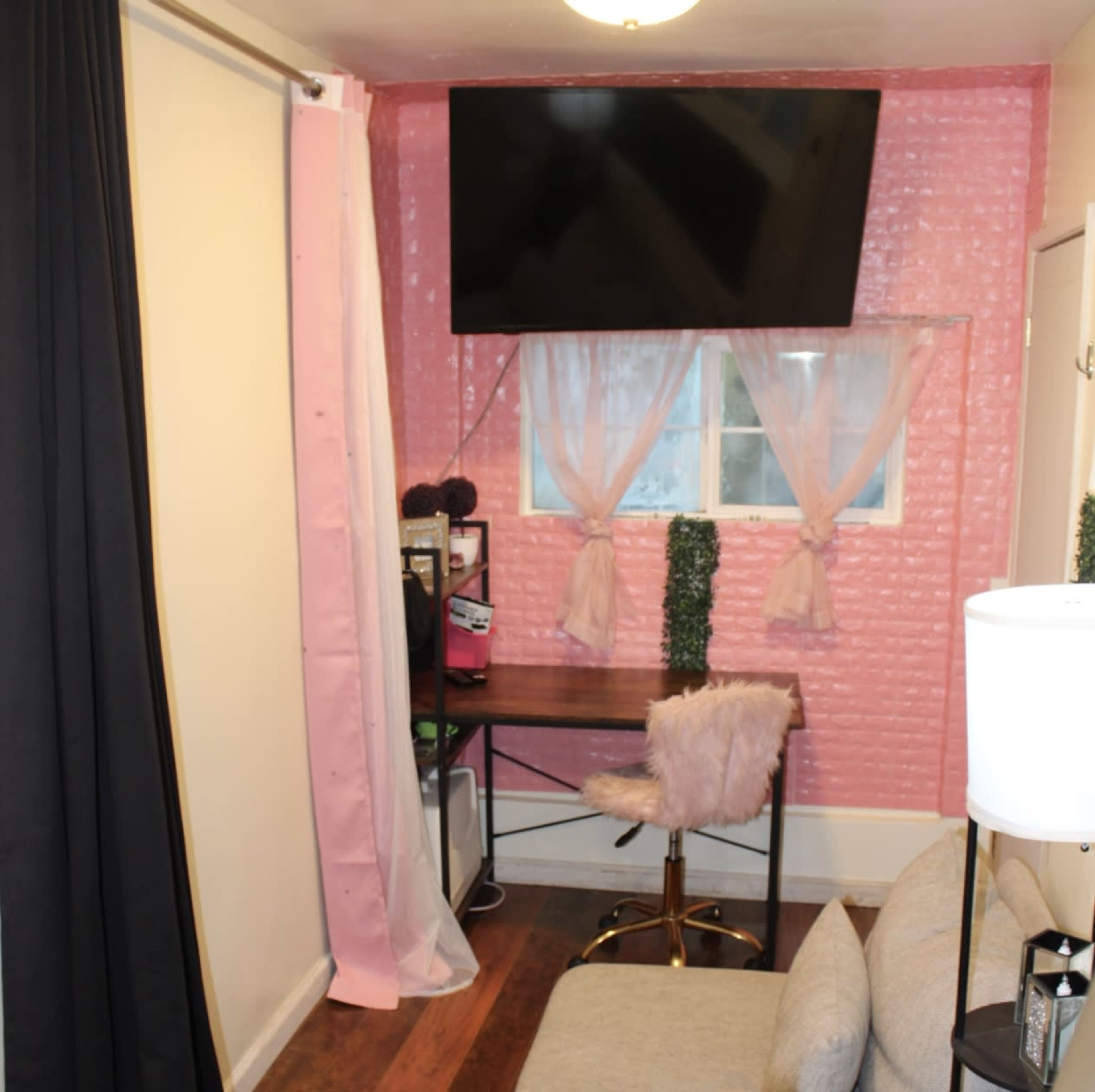 A small room featuring a desk with a gold chair, a large television mounted on a wall, and pink curtains framing a window.