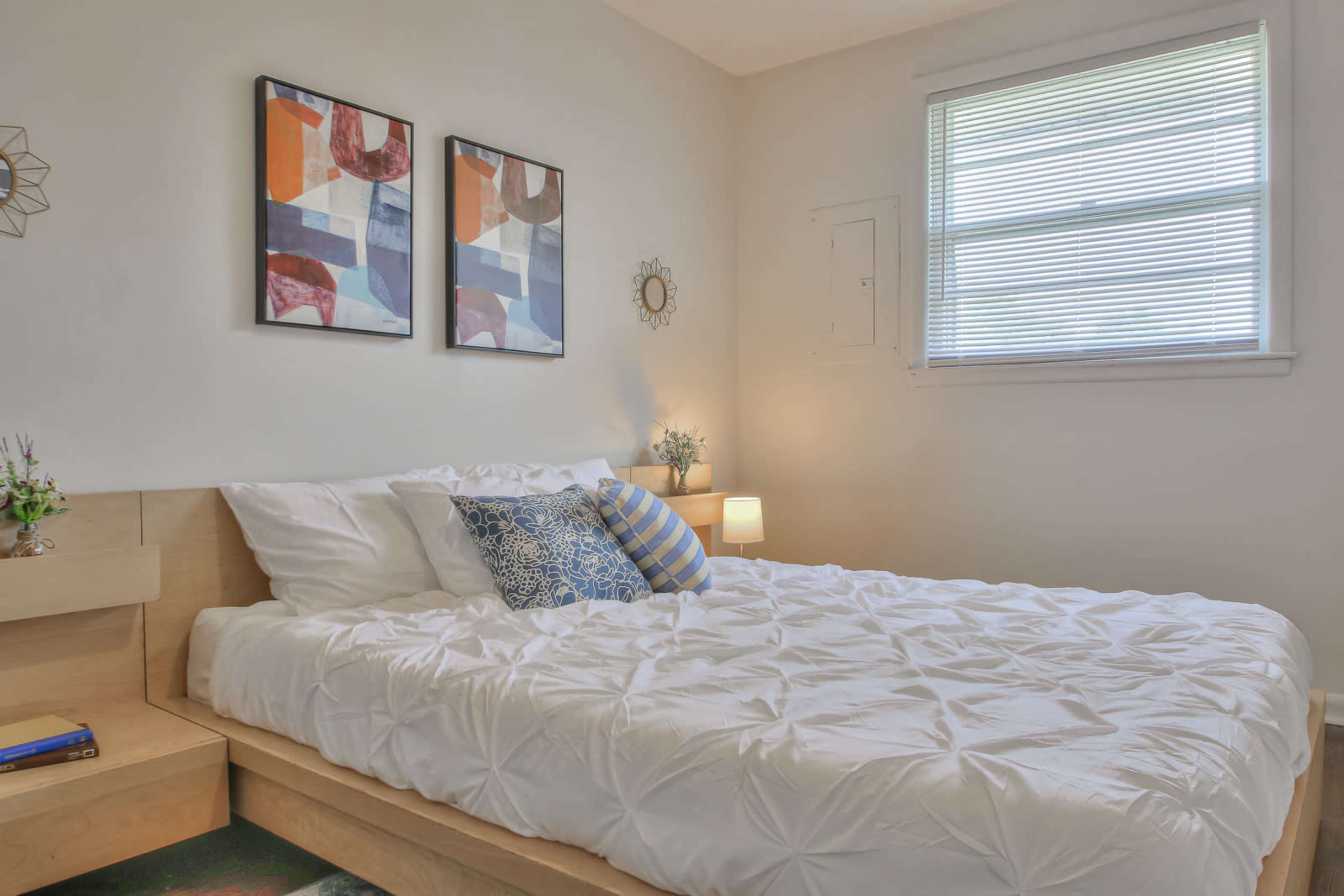 A neatly arranged bedroom features a double bed with white bedding, two framed abstract paintings on the wall, and a small nightstand with a lamp.