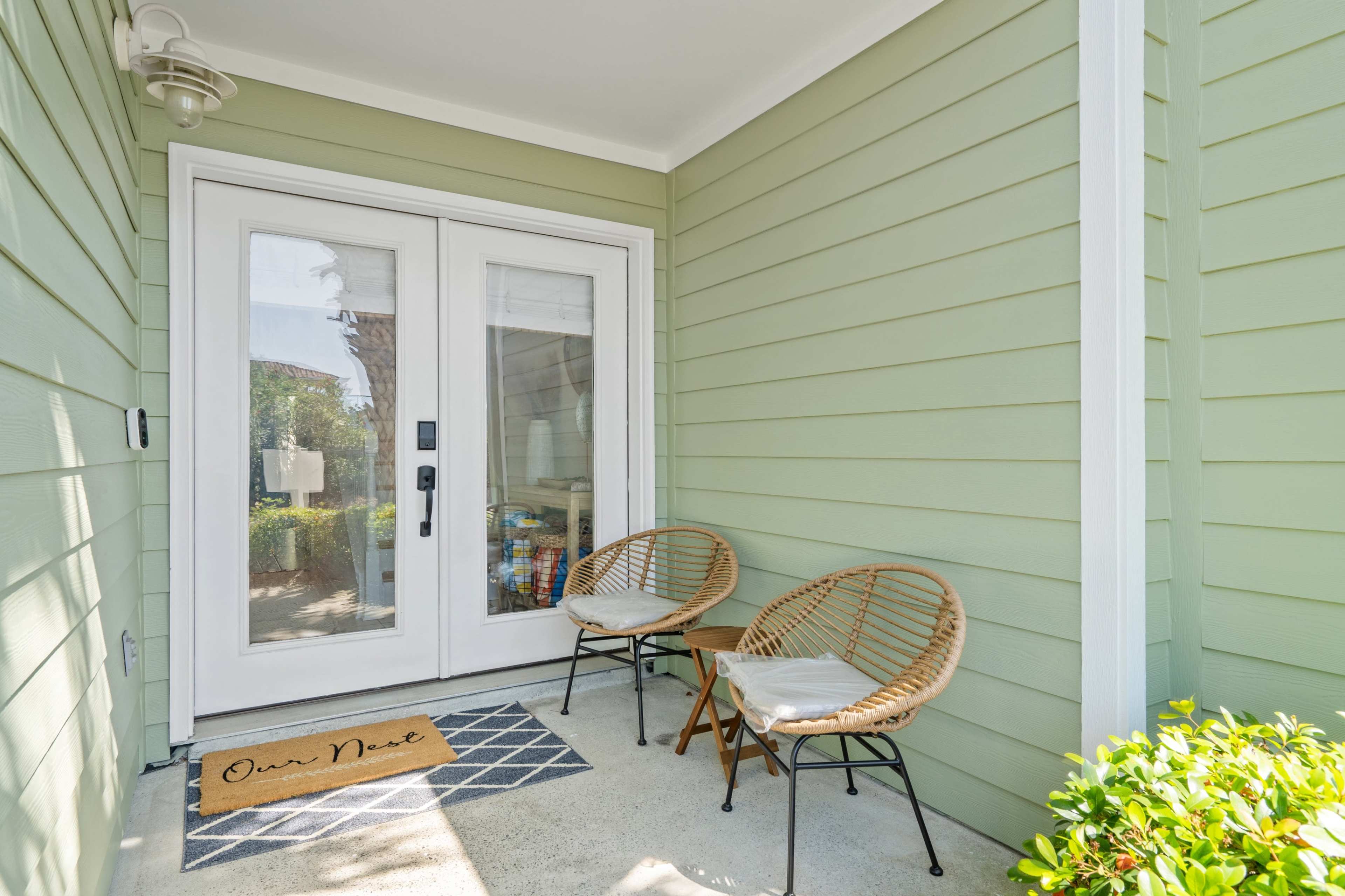 A front porch features two wicker chairs and a welcome mat in front of double glass doors set against a pale green wall.