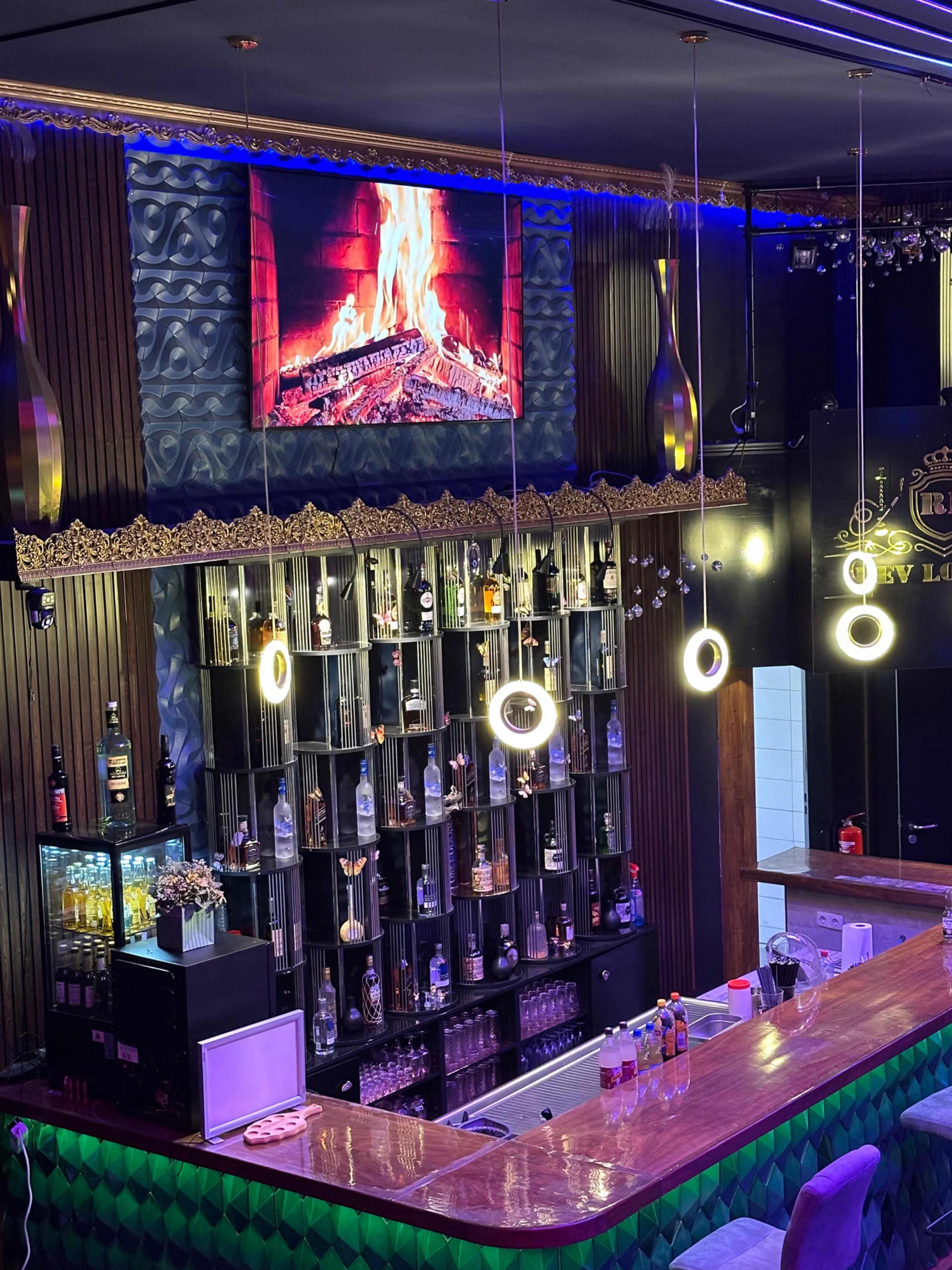 The image shows a modern bar with a well-stocked back shelf, dim lighting, and a television displaying a fire effect.