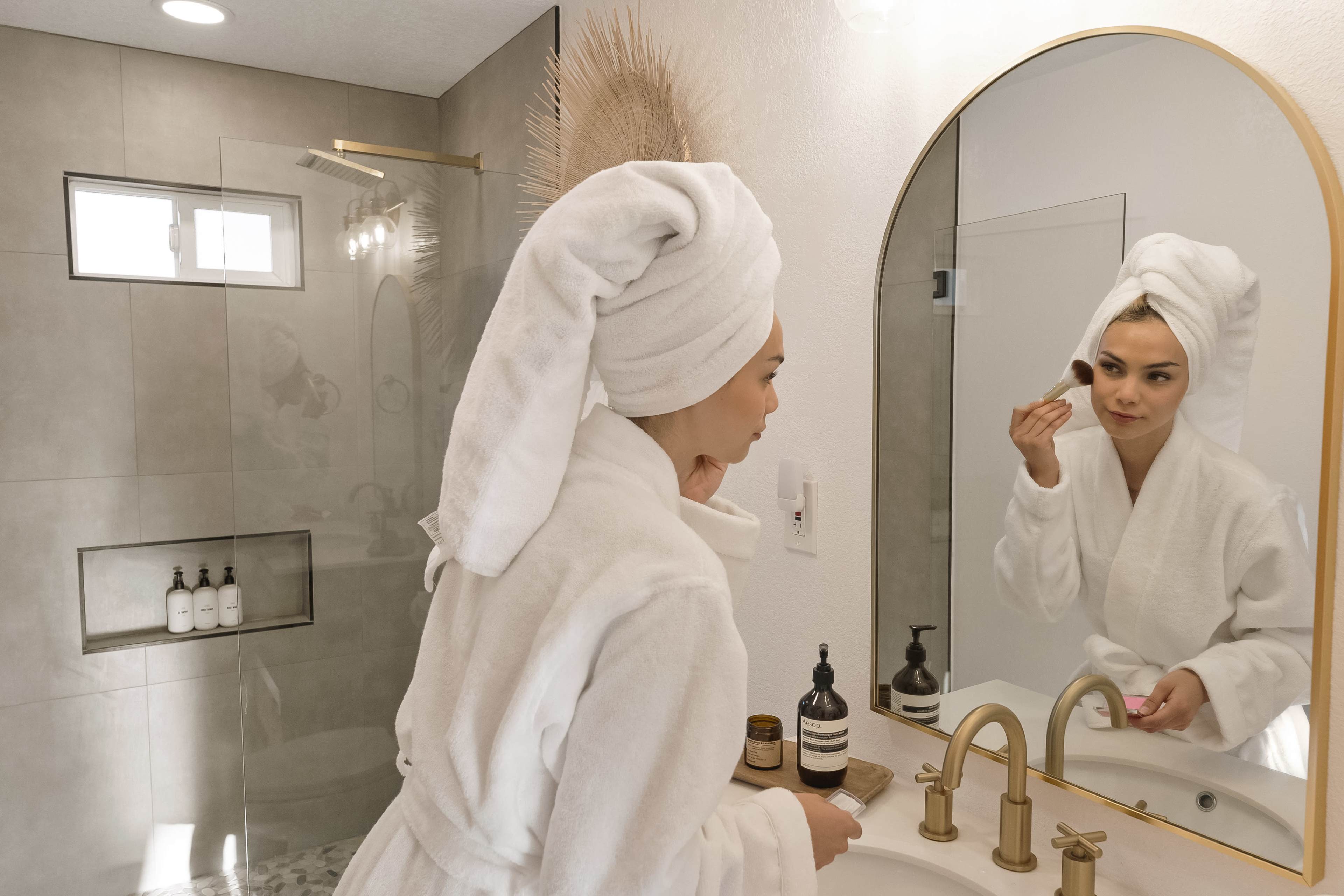 A woman in a white bathrobe applies makeup while looking in a mirror in a modern bathroom.
