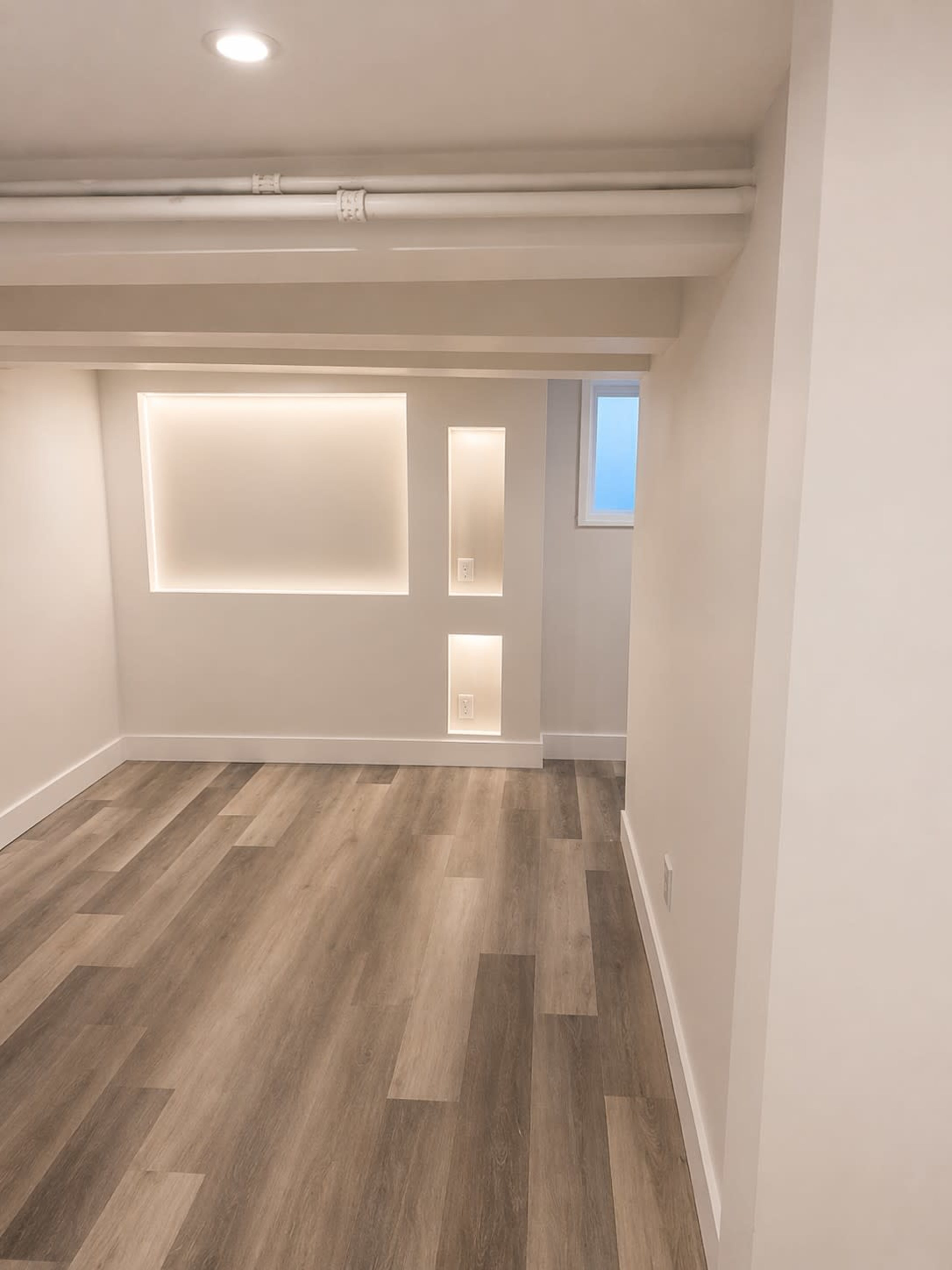 The image shows a modern, unfinished room with light-colored walls and wood flooring, featuring recessed lighting niches on one wall.