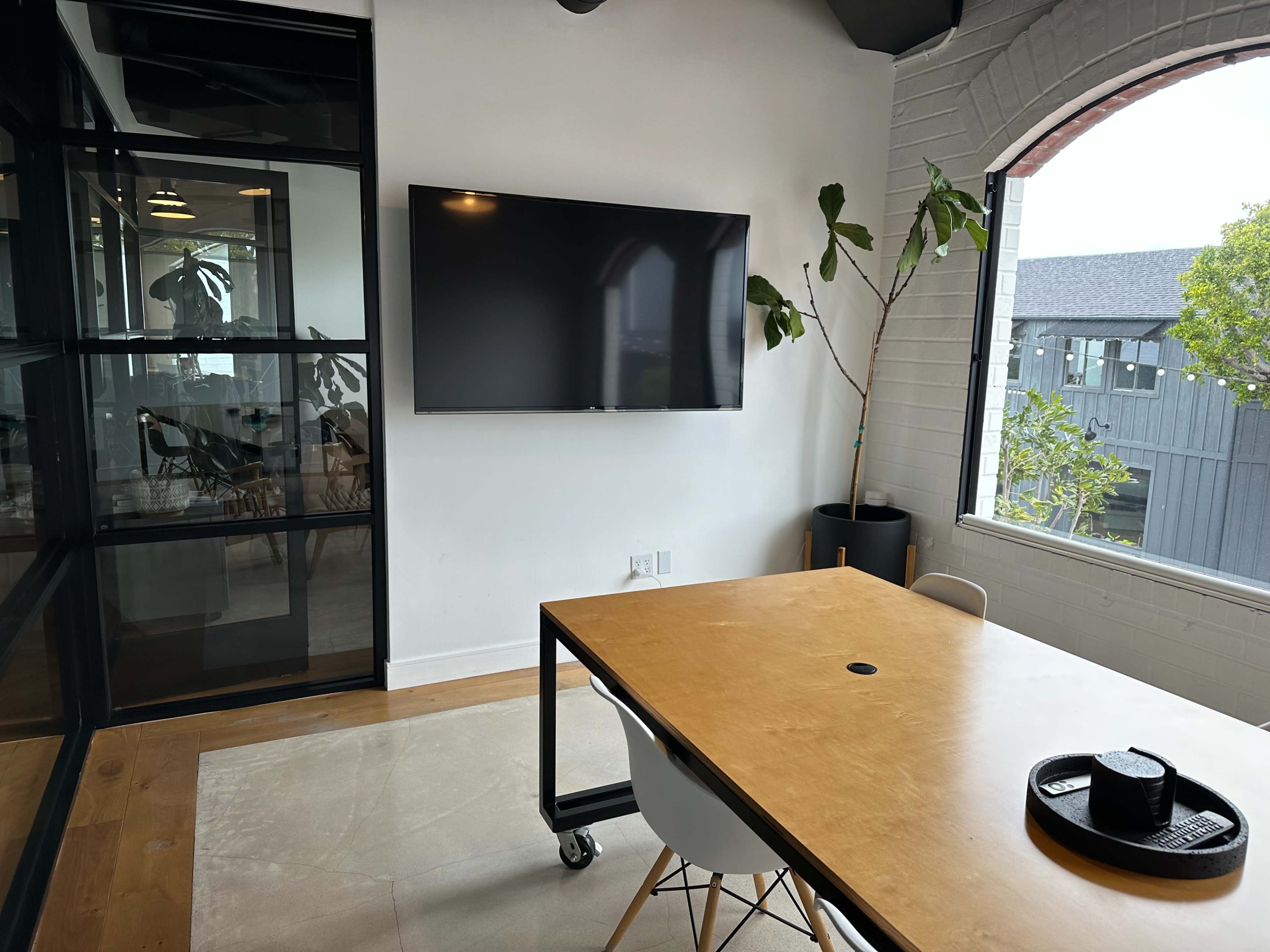 A modern meeting room features a wooden table, a large wall-mounted TV, and a potted plant by the window, with glass walls providing visibility to an adjoining space.