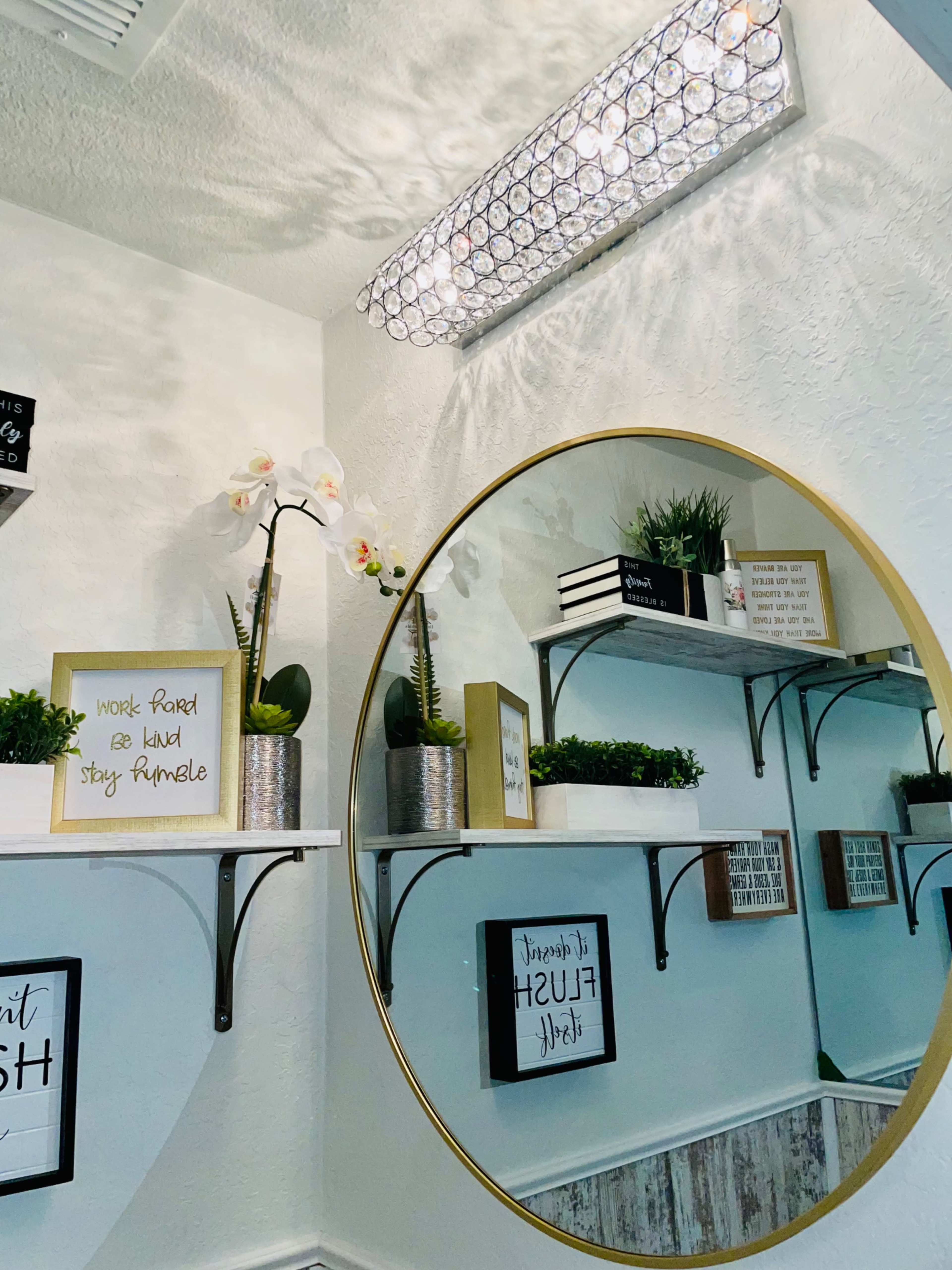 A well-decorated wall with a large circular mirror, shelves featuring plants and decorative signs, and a modern light fixture above.