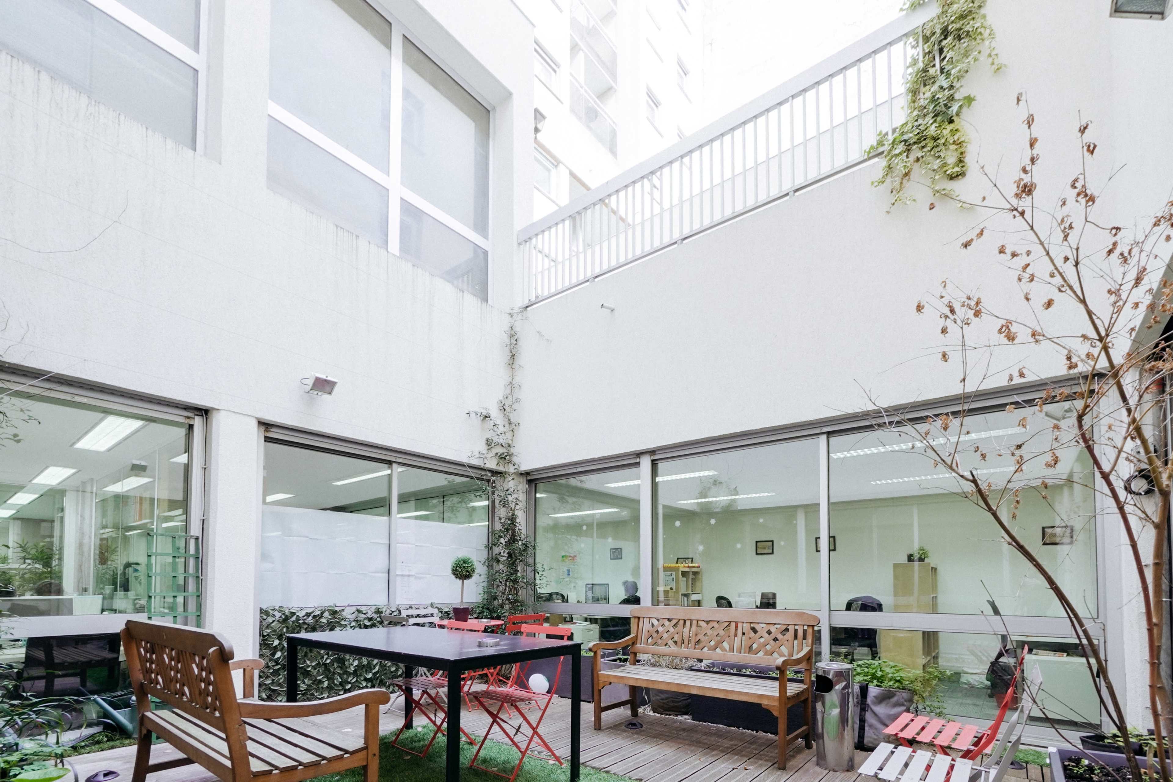 A courtyard features several benches and a table, surrounded by glass walls and greenery.
