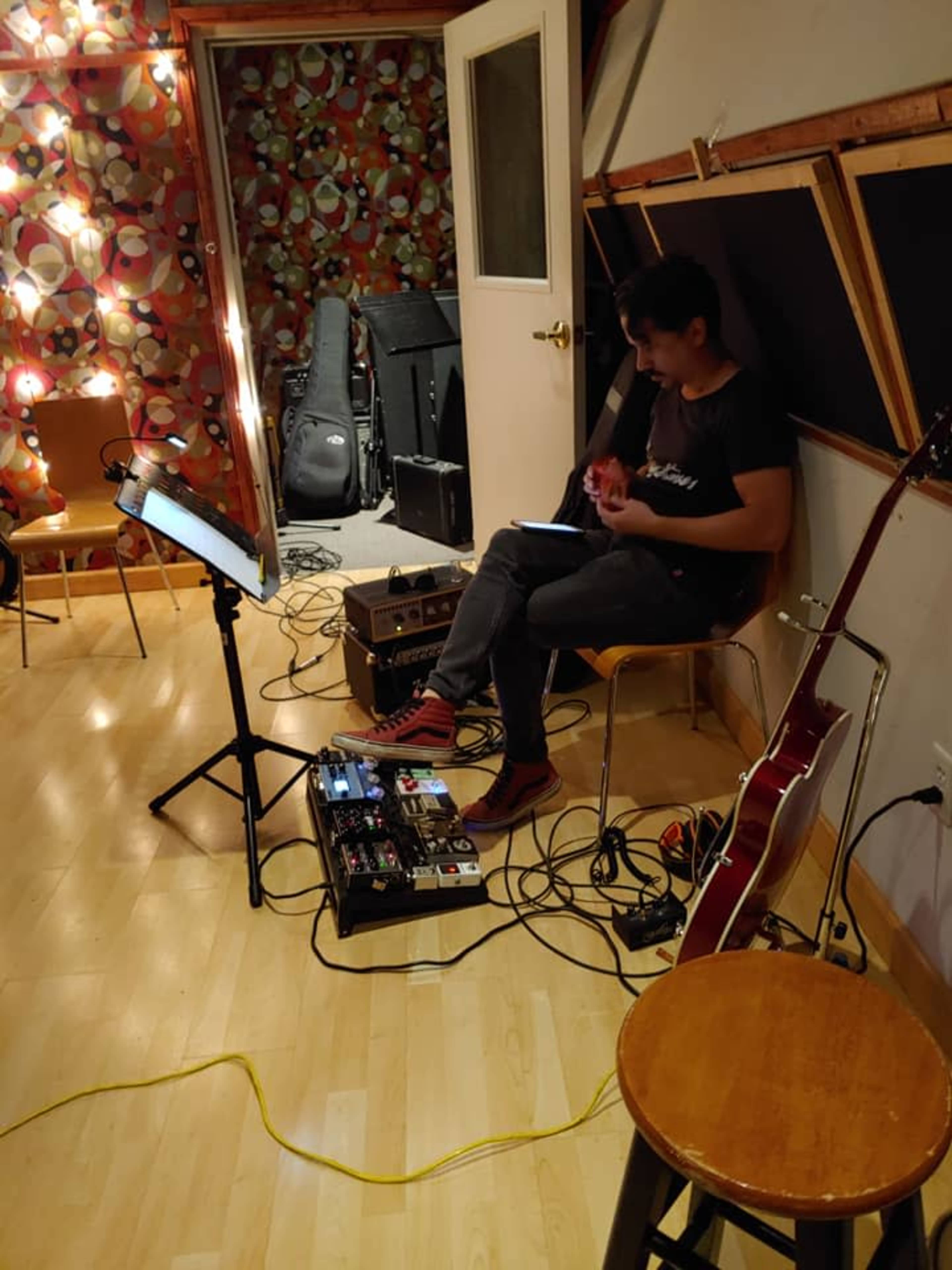A musician sits on a chair in a recording studio, surrounded by instruments and equipment, while playing a guitar.