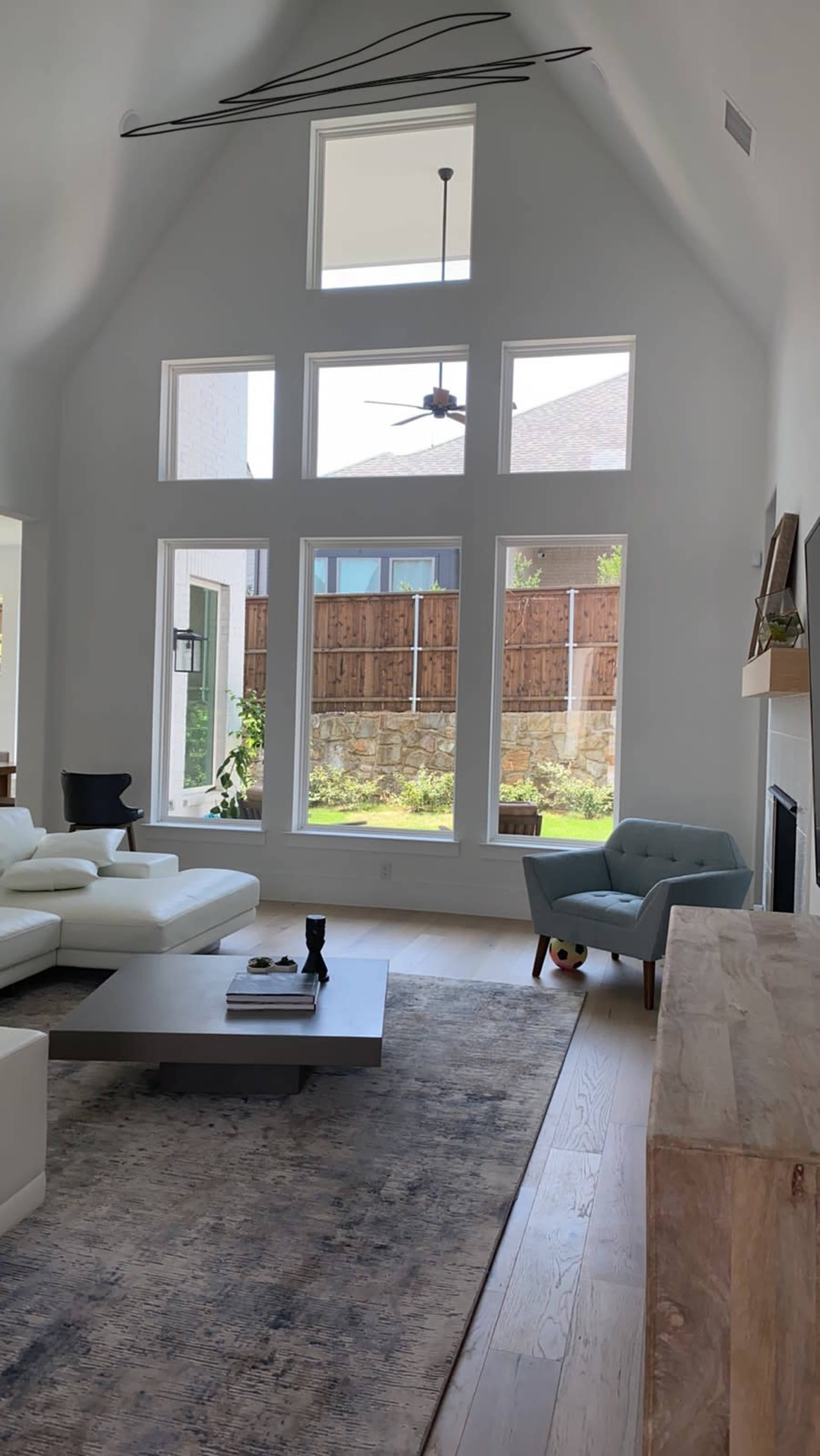 The room features a high ceiling with large windows, a white sectional sofa, a coffee table, and a light blue armchair, overlooking a fenced backyard.