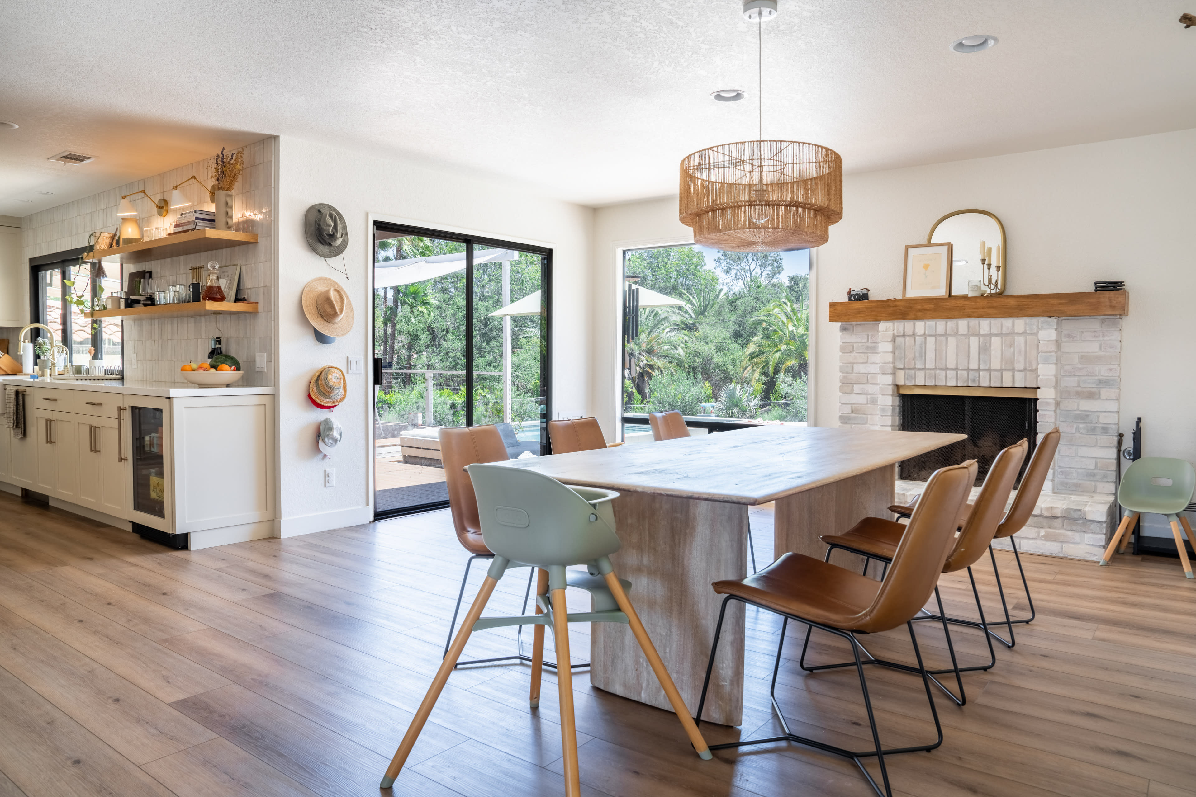 The image shows a modern dining area with a large wooden table surrounded by brown chairs, a fireplace, and large glass doors that open to an outdoor patio.
