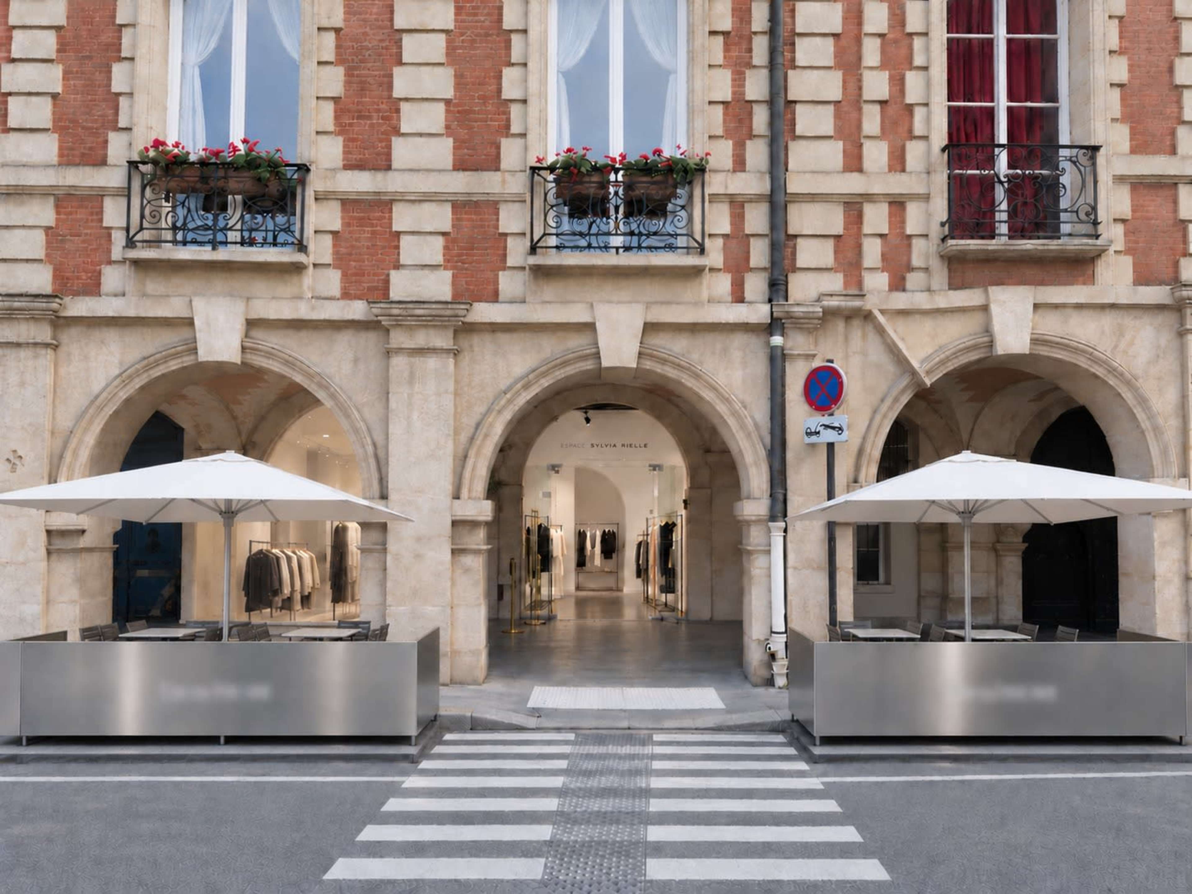 The image shows a clothing store entrance with large glass windows and two outdoor seating areas beneath white umbrellas, situated in a brick building with arched doorways.