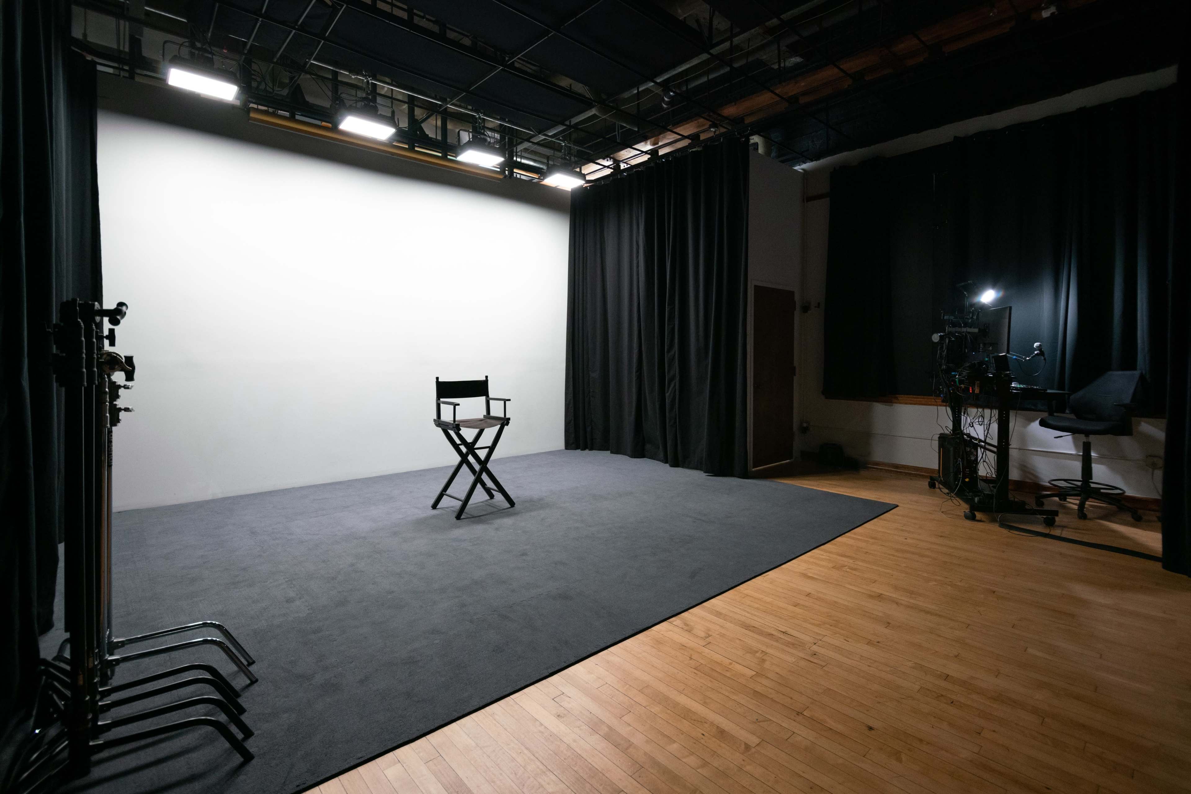 A spacious, empty studio with a single director's chair facing a blank wall, surrounded by black curtains and dim lighting.