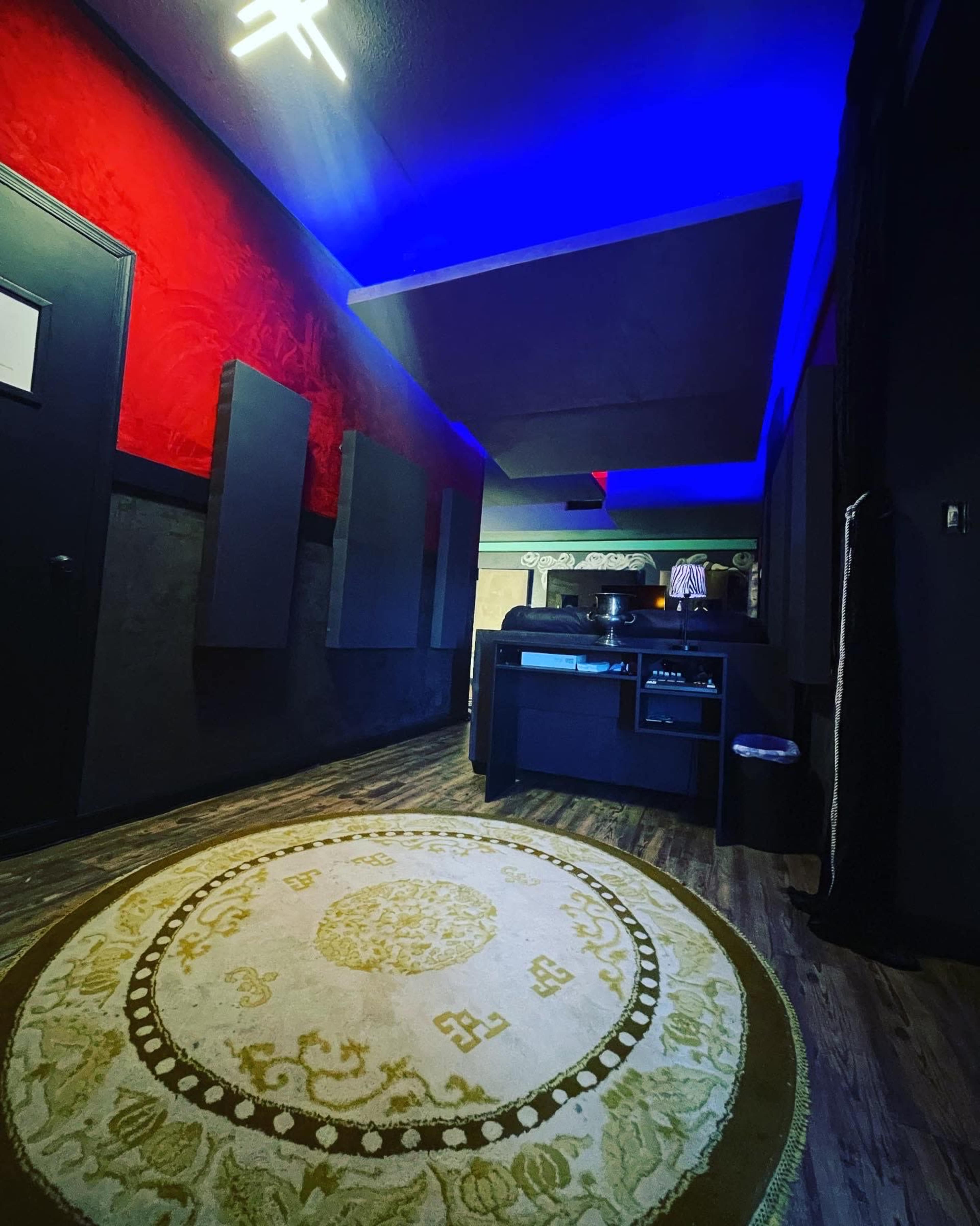 A room with a patterned rug, dark walls, padded panels, and blue lighting, featuring a workstation at the far end.