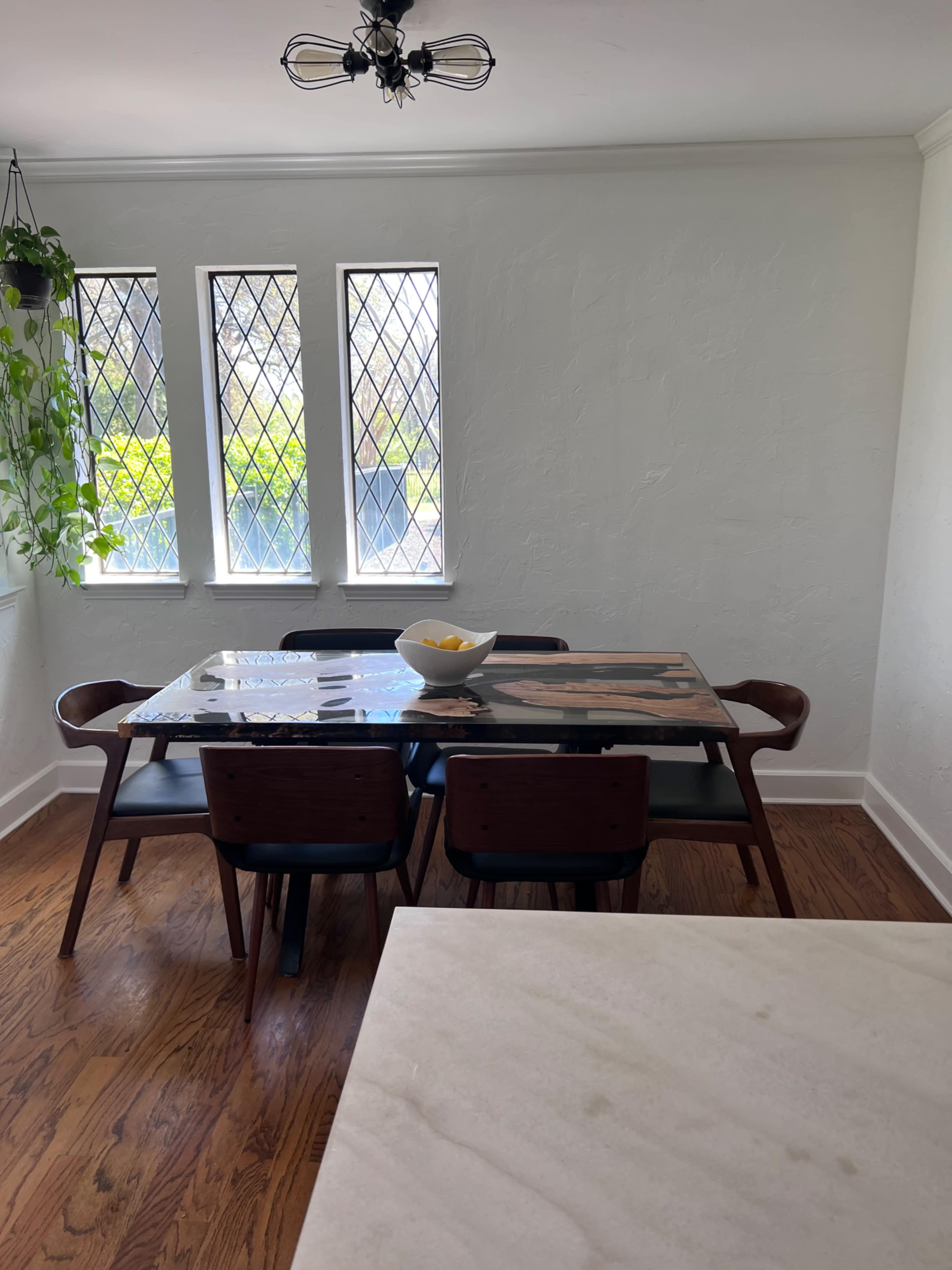 A dining area features a wooden table surrounded by chairs, with large diamond-patterned windows and a potted plant in the corner.