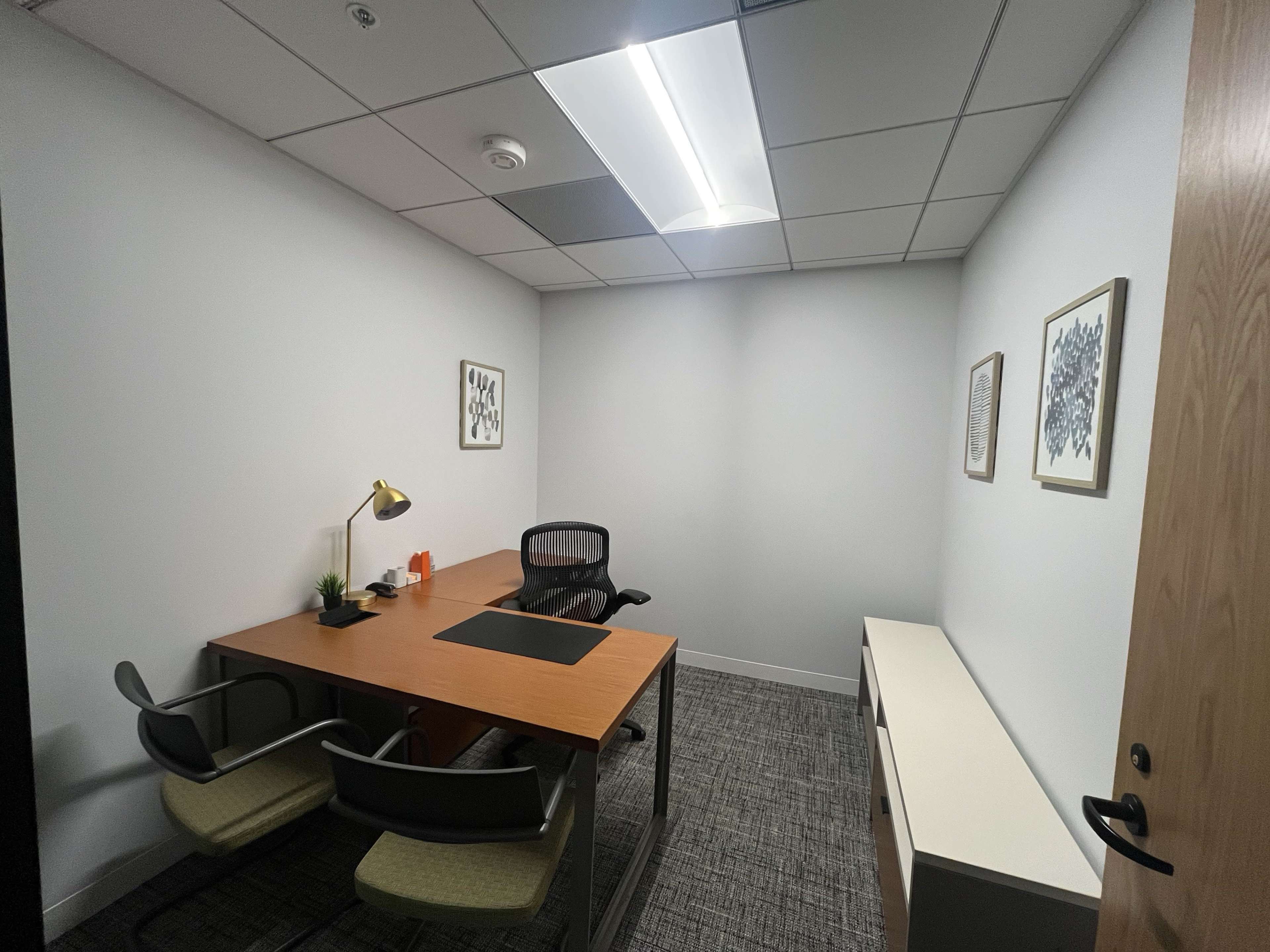 The image shows a small, modern office space with a desk, two chairs, and minimalist decor on the walls.