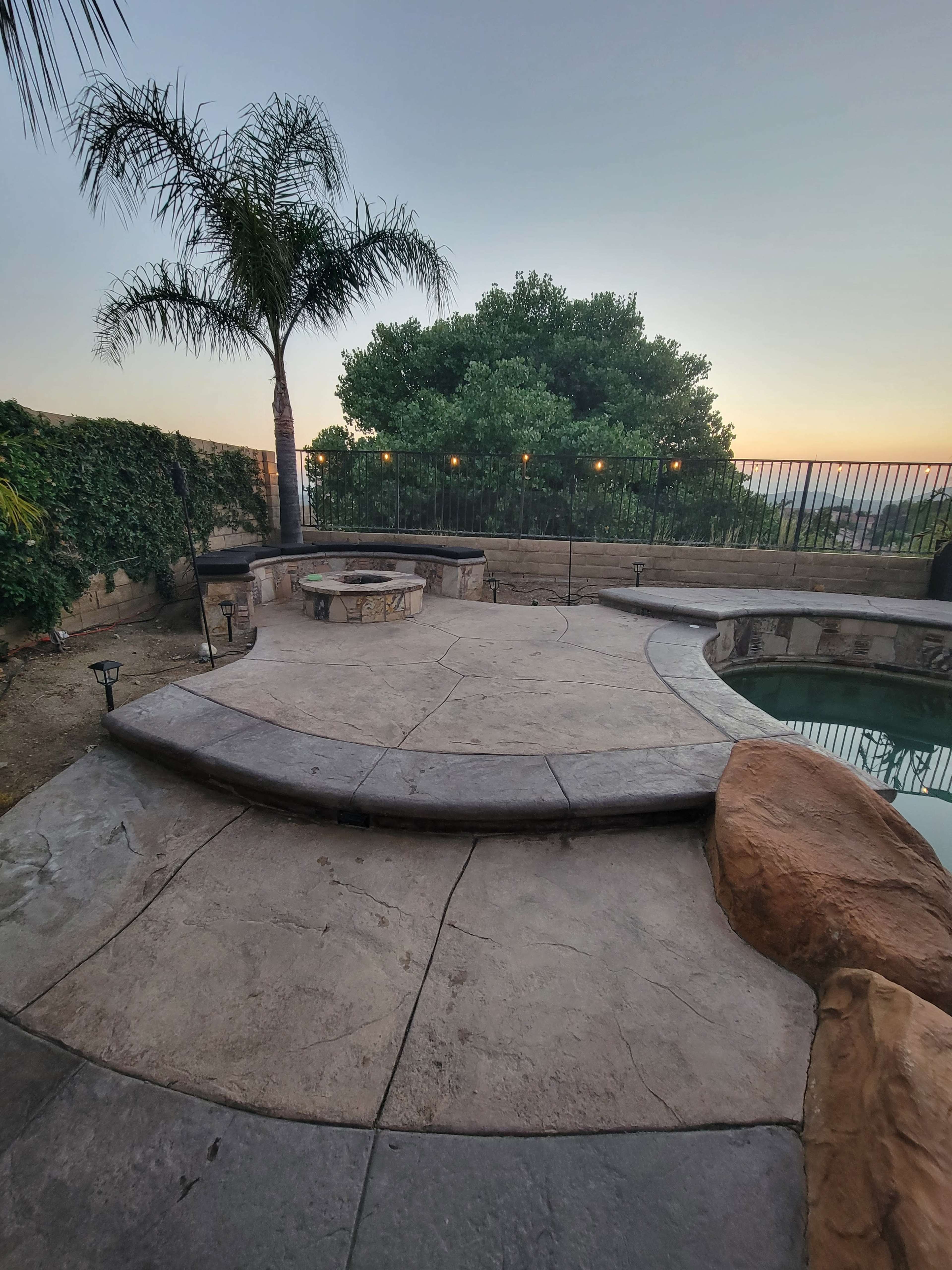 A landscaped outdoor area featuring a stone patio, a round seating arrangement, a swimming pool, and palm trees against a sunset sky.