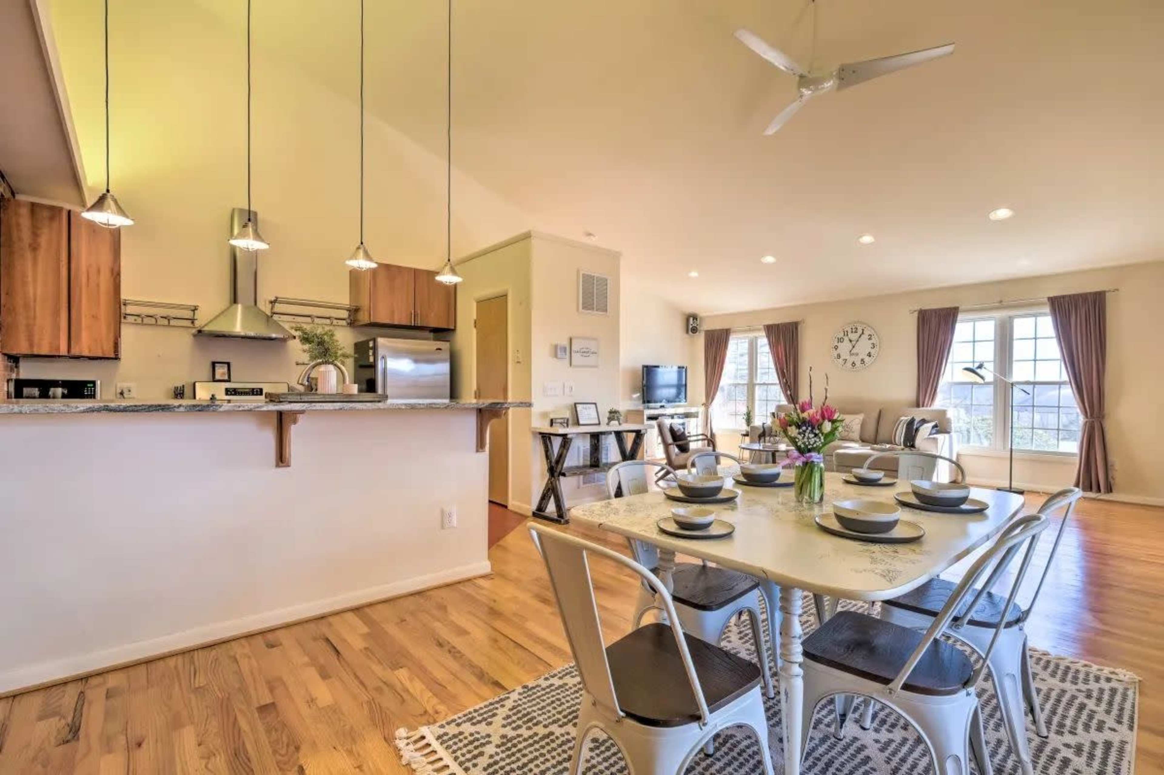 A modern dining area features a table set for six, surrounded by metal chairs, adjacent to an open kitchen with wooden cabinets and stainless steel appliances.