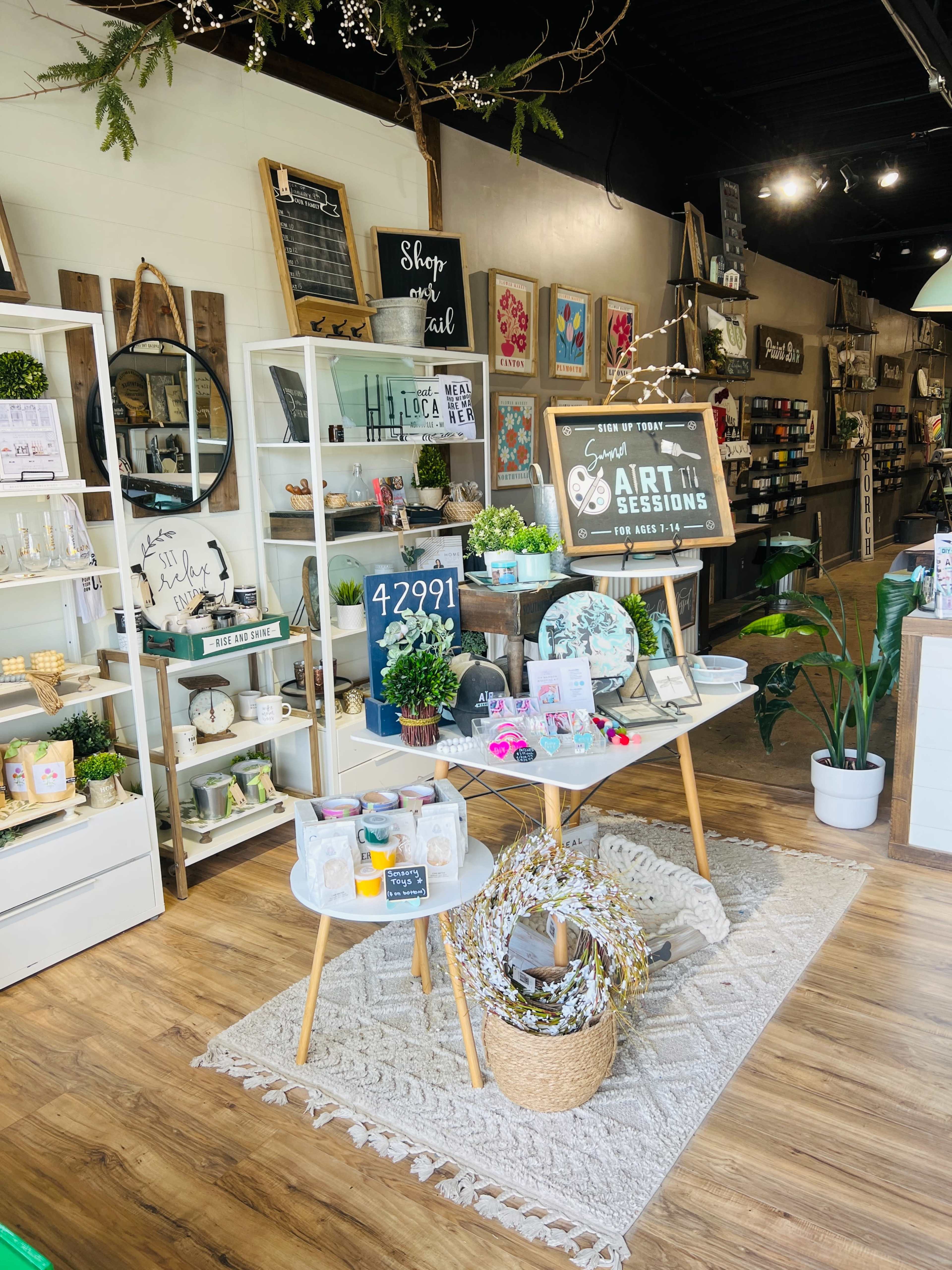 A bright boutique interior featuring display shelves filled with various decor items and a central table showcasing a sign for art sessions surrounded by plants and seasonal decorations.