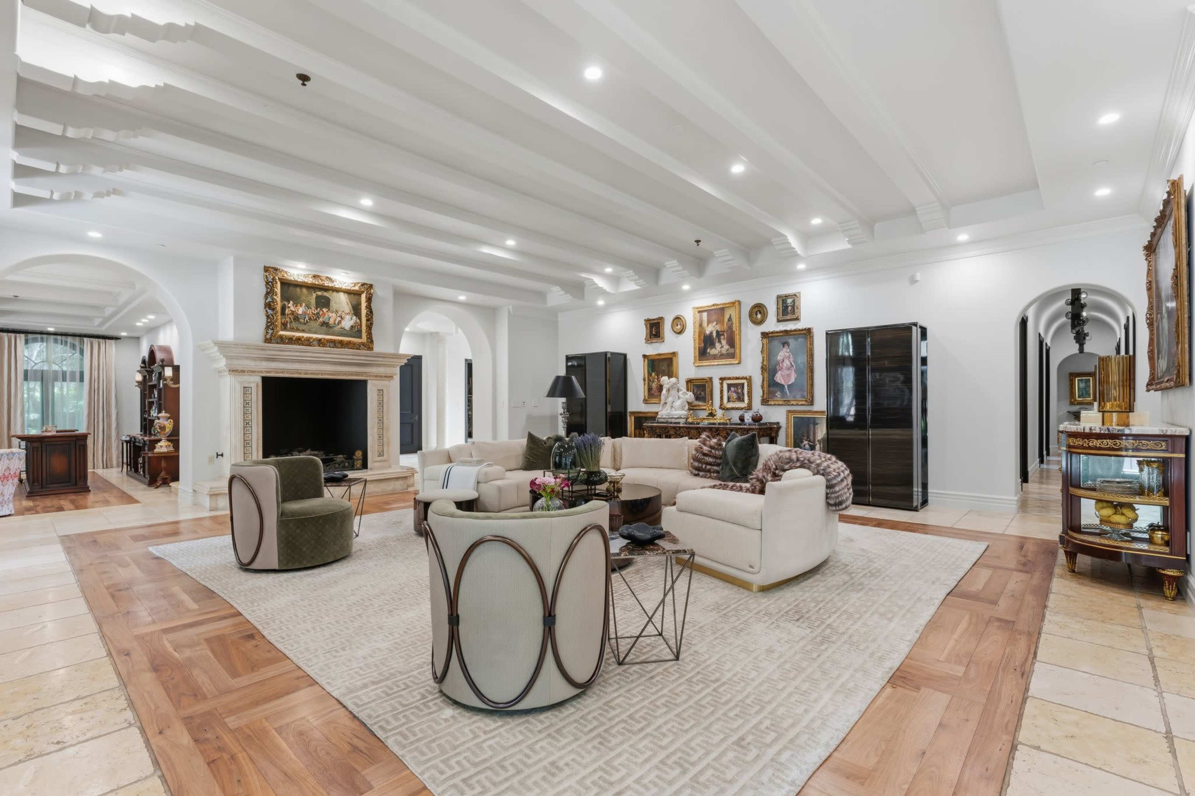 The image features a spacious living room with a beige sectional sofa, round chairs, a large area rug, and a fireplace, adorned with various artworks and decorative items.